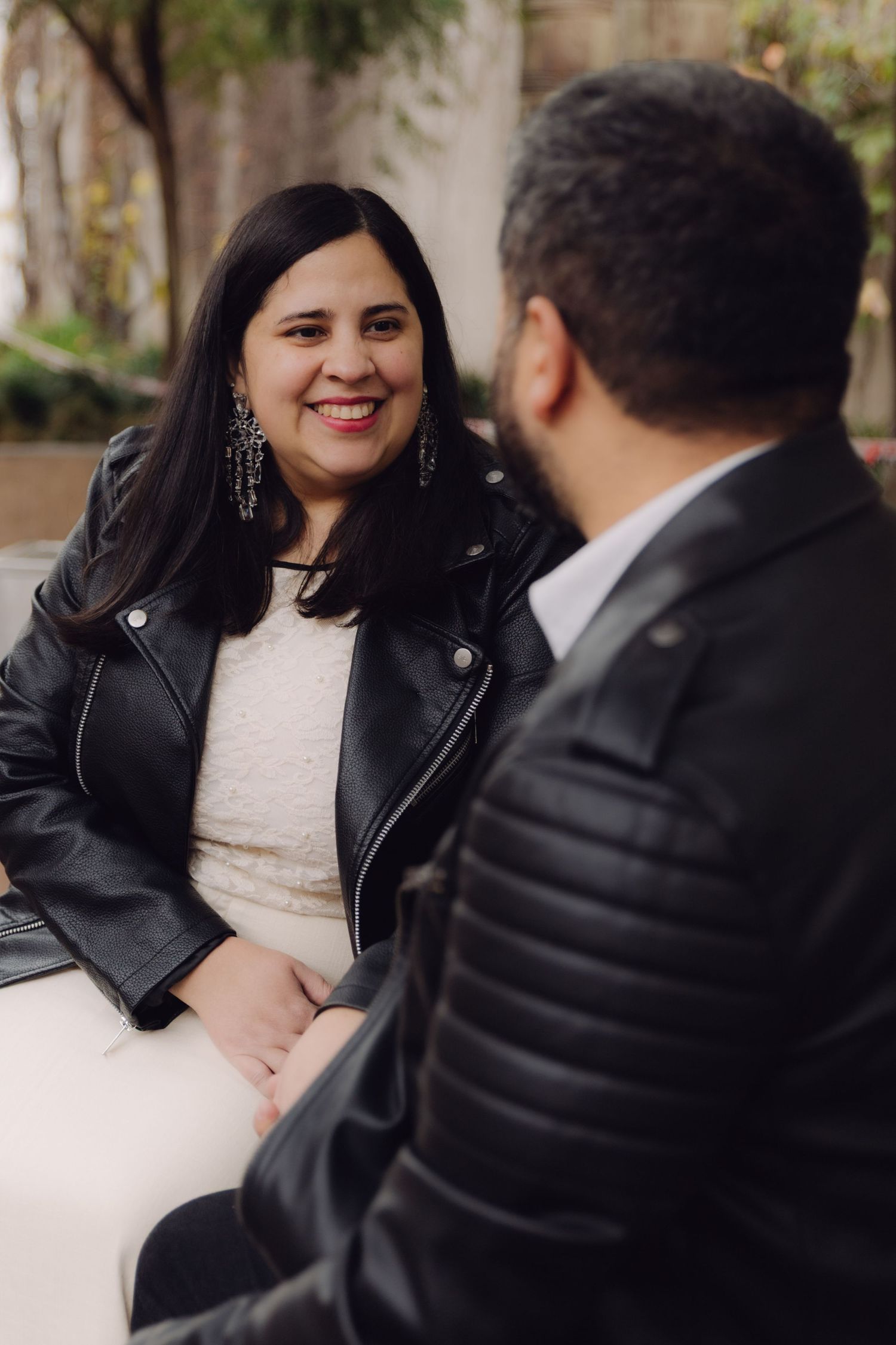 Smiling woman in white dress and black leather jacket holding hands with man during outdoor couples session.