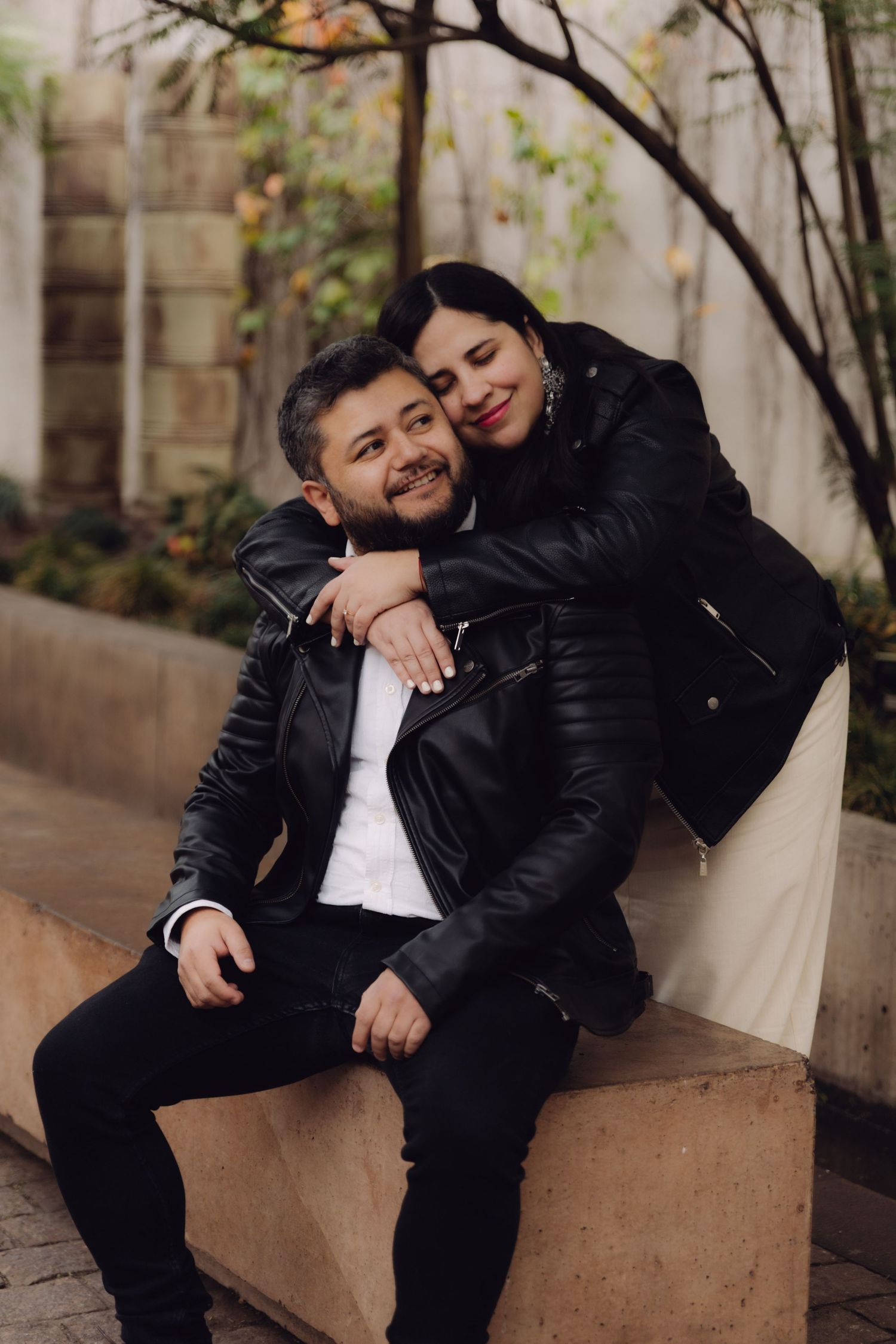 Affectionate couple in black leather jackets embracing on a stone bench with urban greenery background.