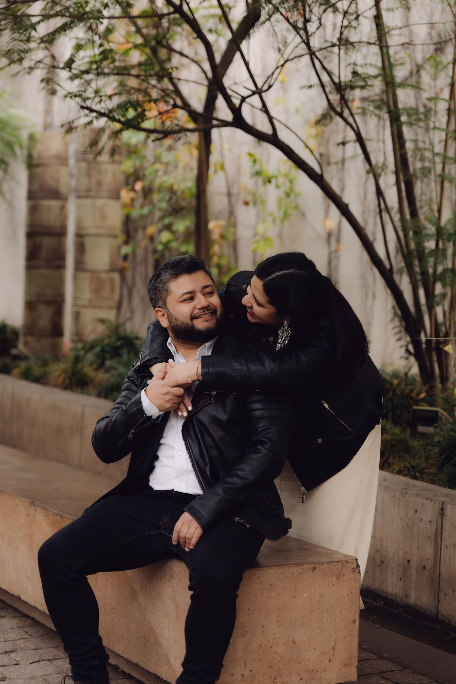 Happy couple in leather jackets sharing a playful moment on an outdoor stone bench surrounded by trees.