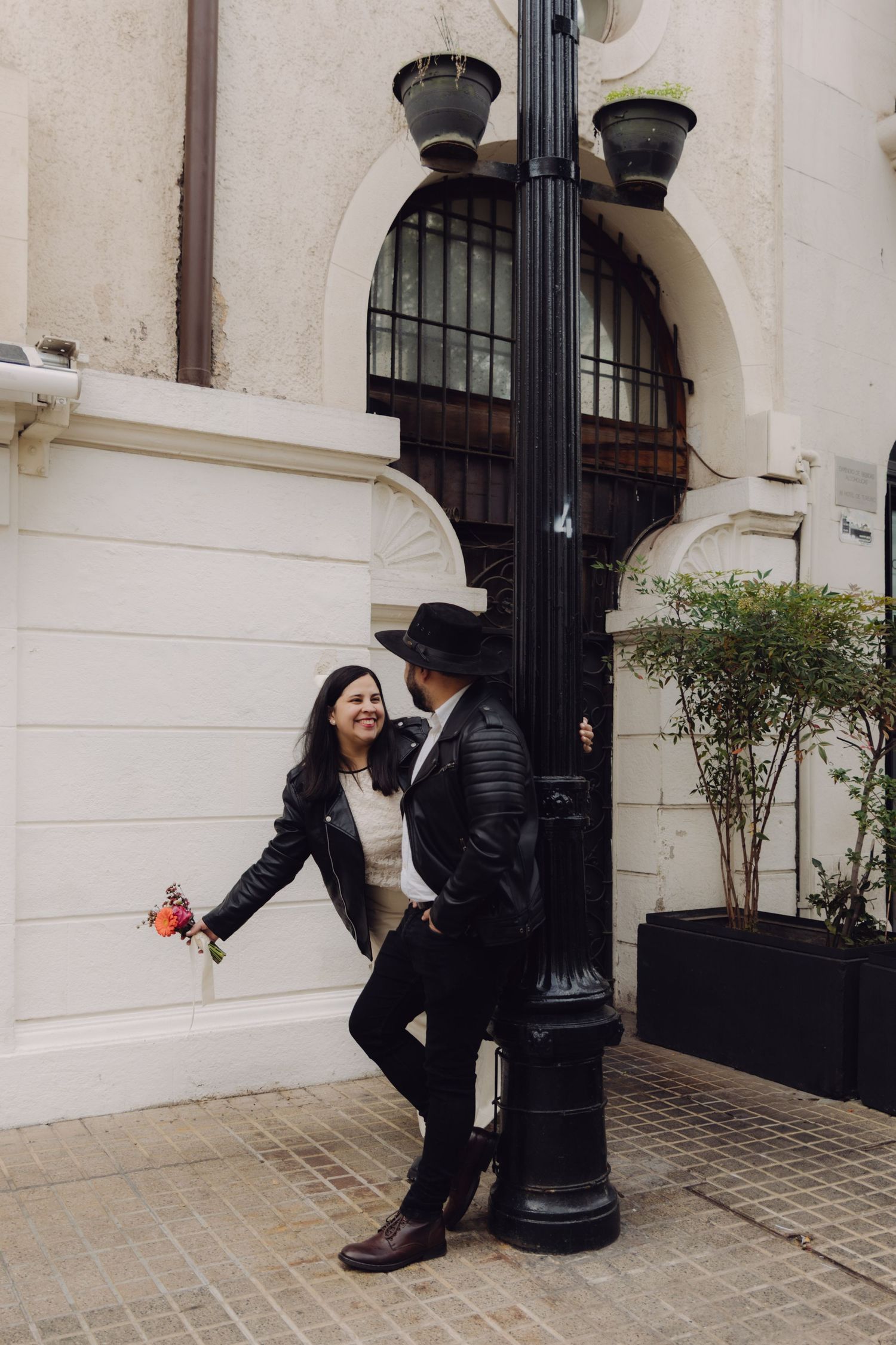 Couple in black leather jackets smiling at each other beside an ornate street lamp on a city sidewalk.