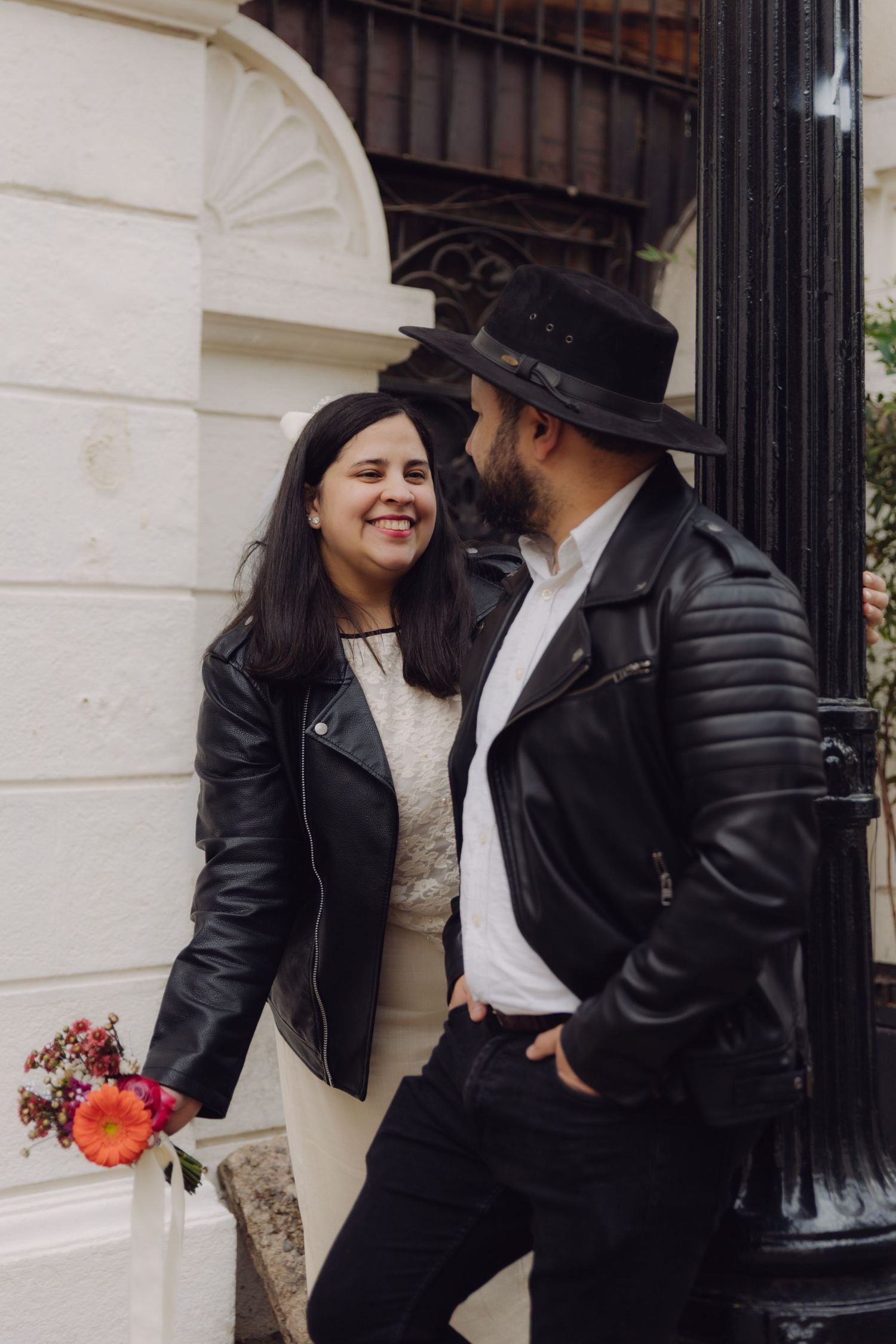 Smiling couple in black leather jackets standing close together near a decorative street lamp with flowers.