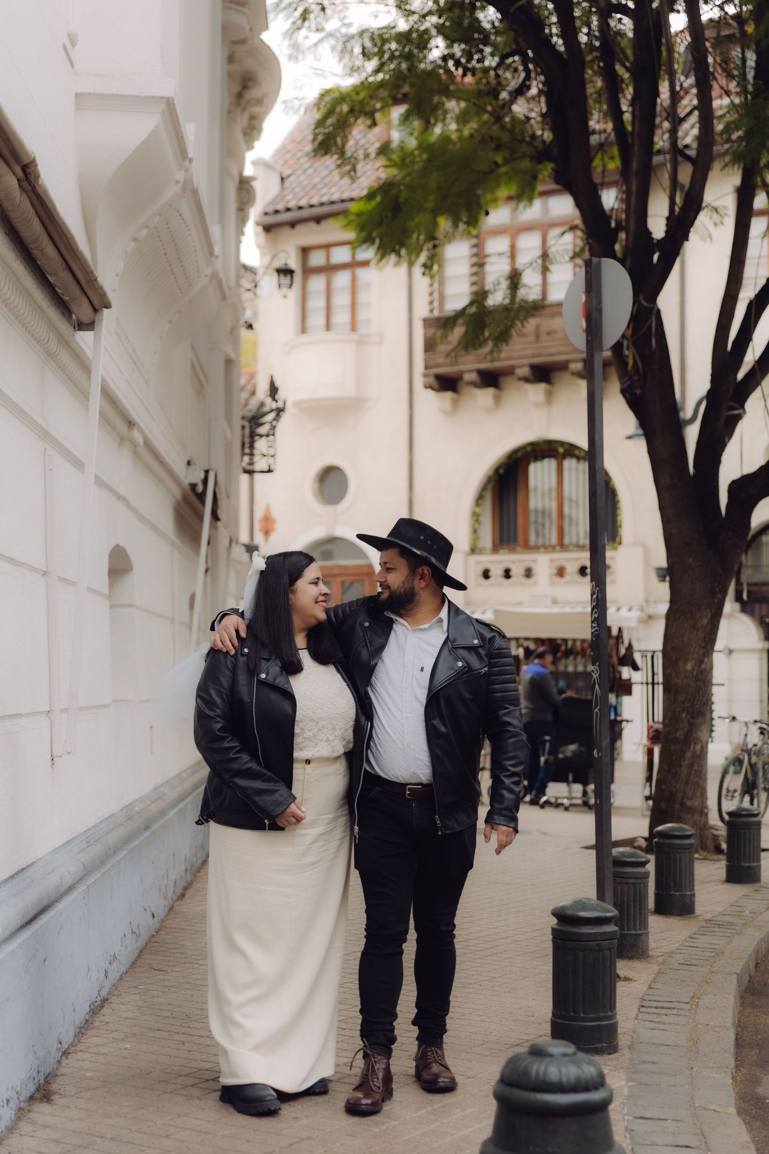 Romantic couple in black leather jackets walking arm in arm down a charming European-style city street.