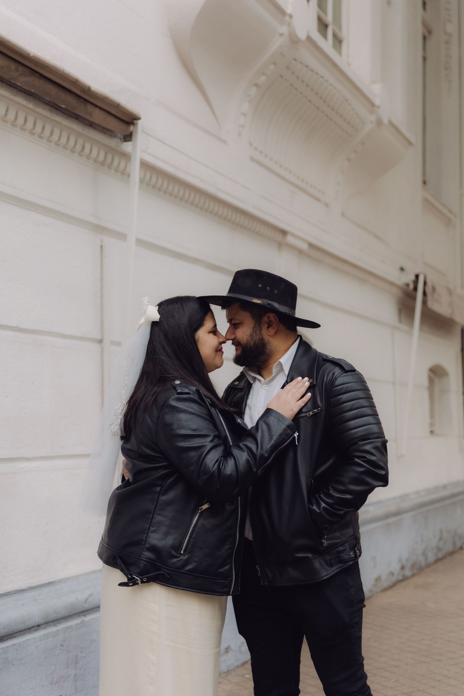 Couple in black leather jackets sharing a romantic almost-kiss moment against a white building exterior.