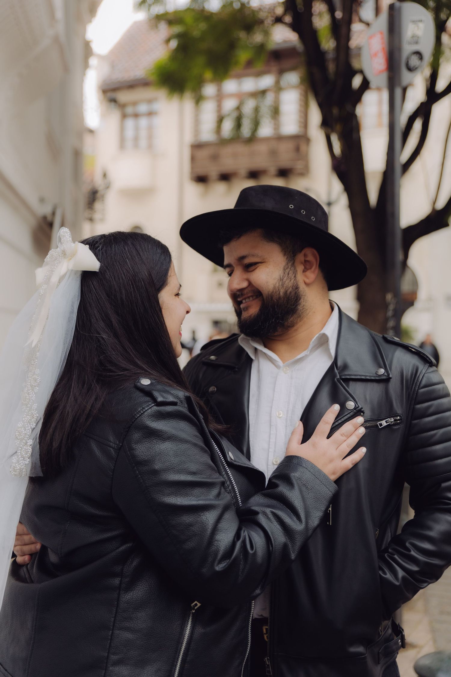 Newlywed couple in black leather jackets smiling at each other on a city street, bride wearing a veil.