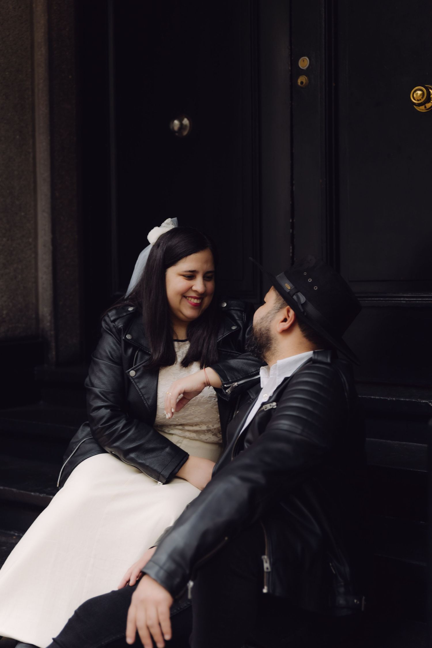 Smiling bride in leather jacket and veil sits on steps with bearded groom in black hat, sharing a tender moment.