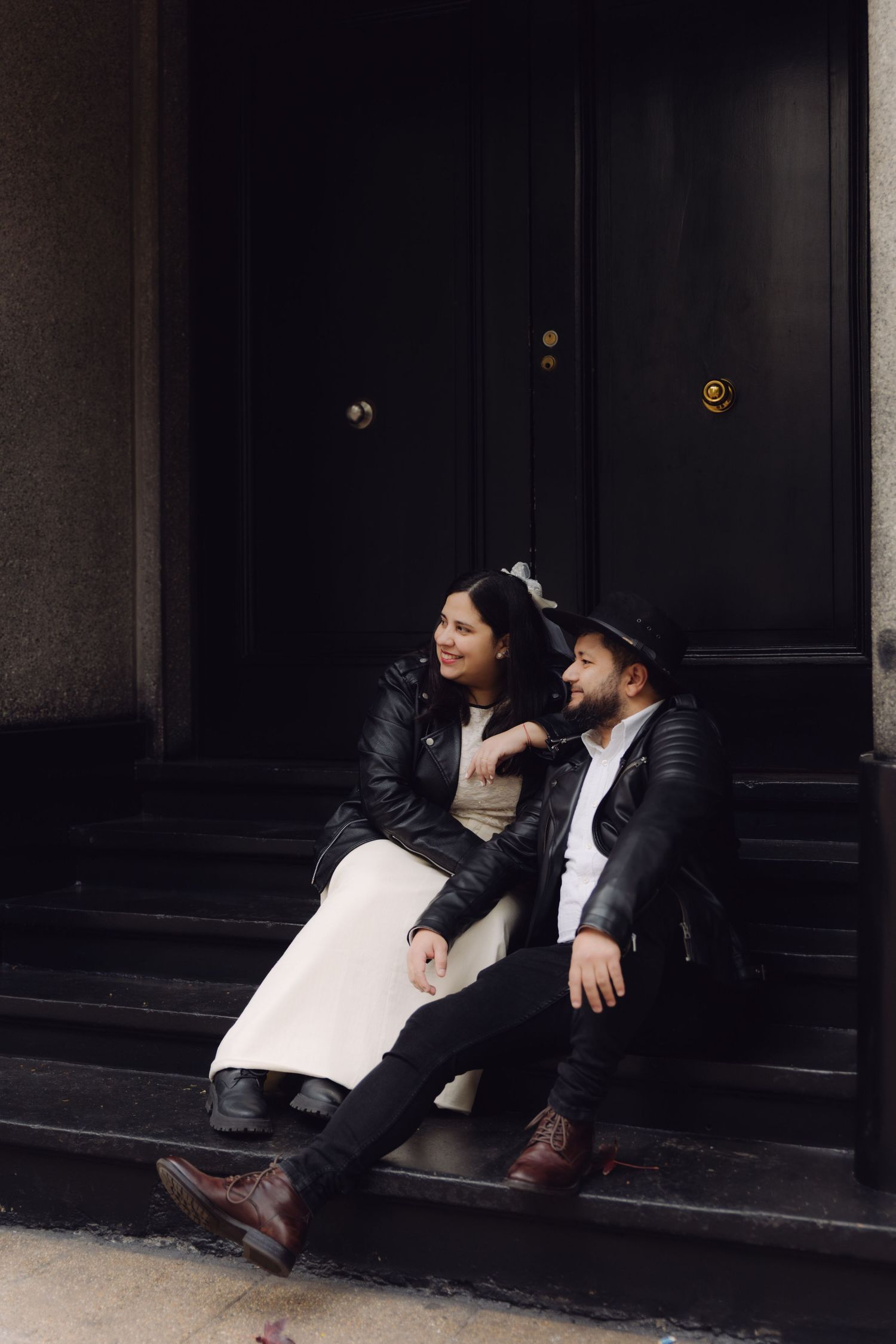 Stylish couple in matching leather jackets sit on dark stone steps outside a building, gazing at each other warmly.