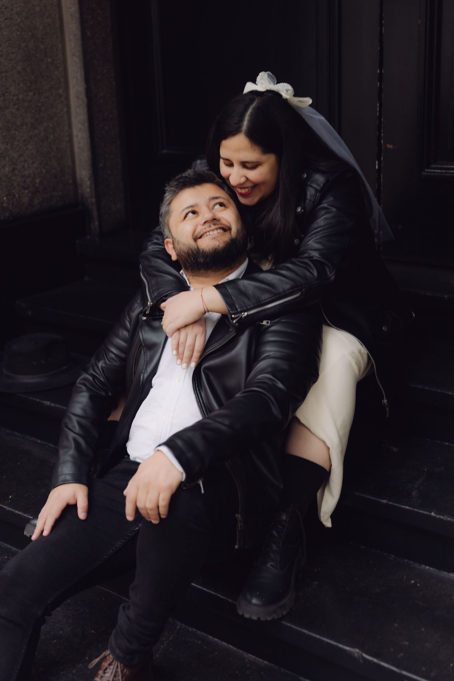 Bride in white dress and leather jacket embraces bearded groom from behind on dark steps, both smiling joyfully.