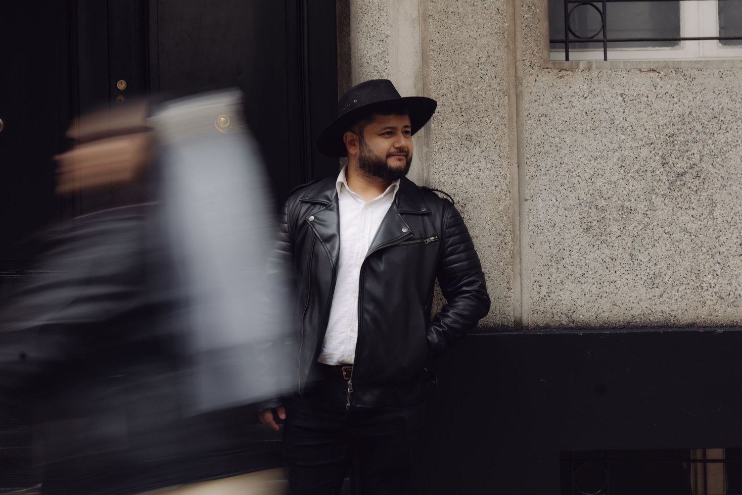 Stylish bearded man in black leather jacket and hat stands confidently outside a building with blurred motion nearby.