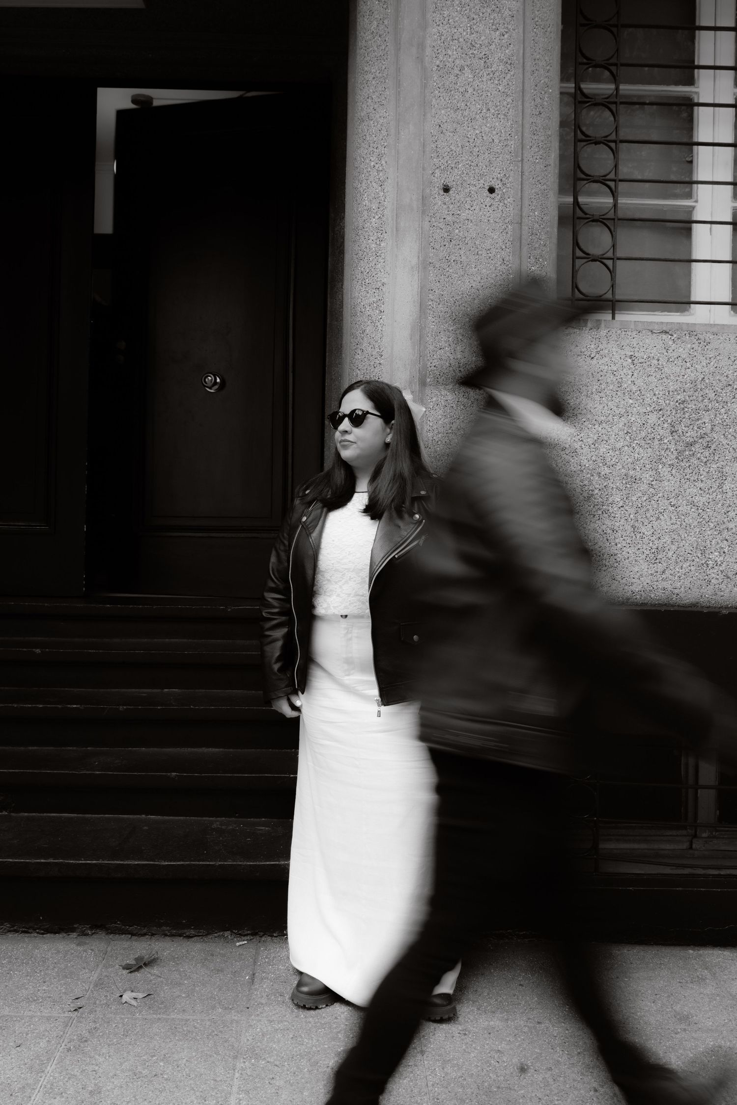 Black and white photo of bride in white dress and leather jacket posing outside building with blurred pedestrian passing.