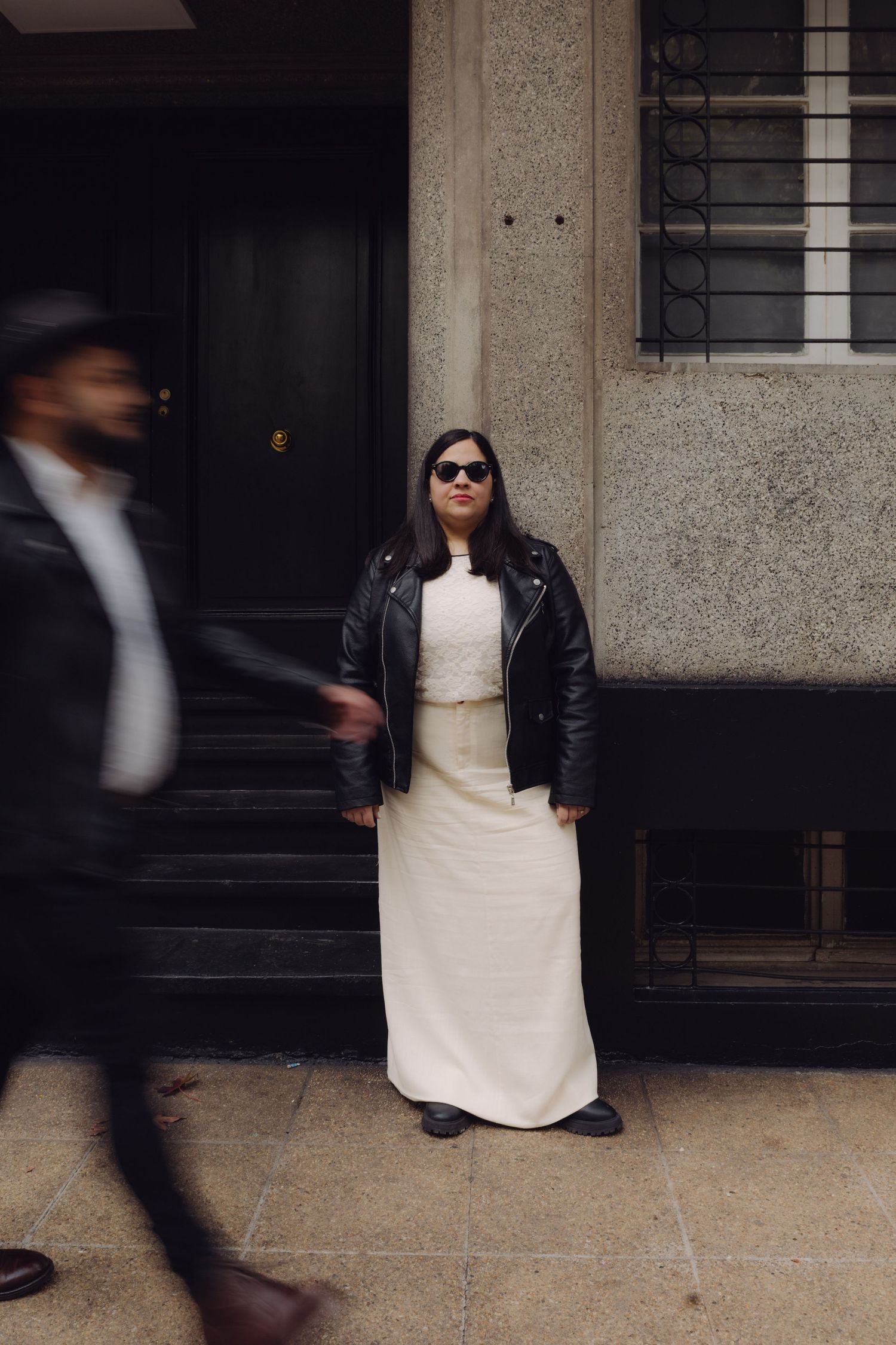 Bride in white maxi dress, leather jacket, and sunglasses stands confidently outside building as blurred figure walks past.