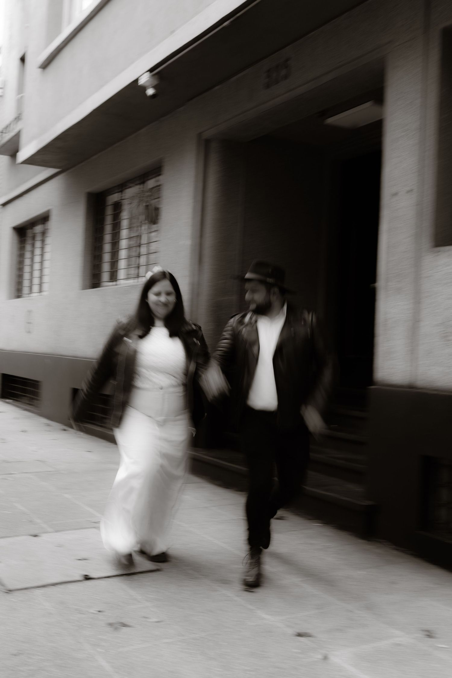 Black and white blurred motion photo of bride and groom in leather jackets walking together outside urban building.