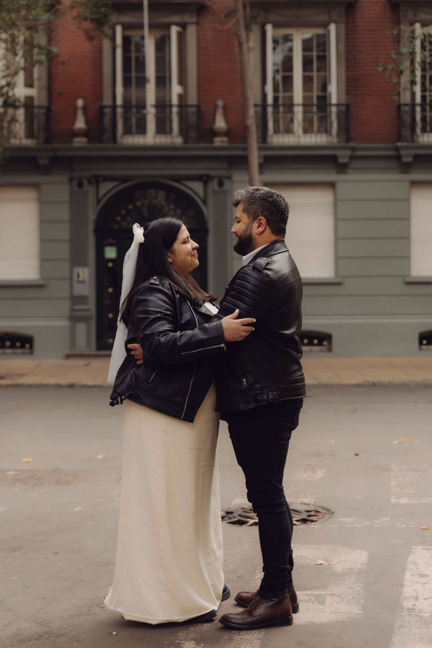 Bride in white dress with veil and groom in black leather jacket embrace face-to-face on a rainy city street.