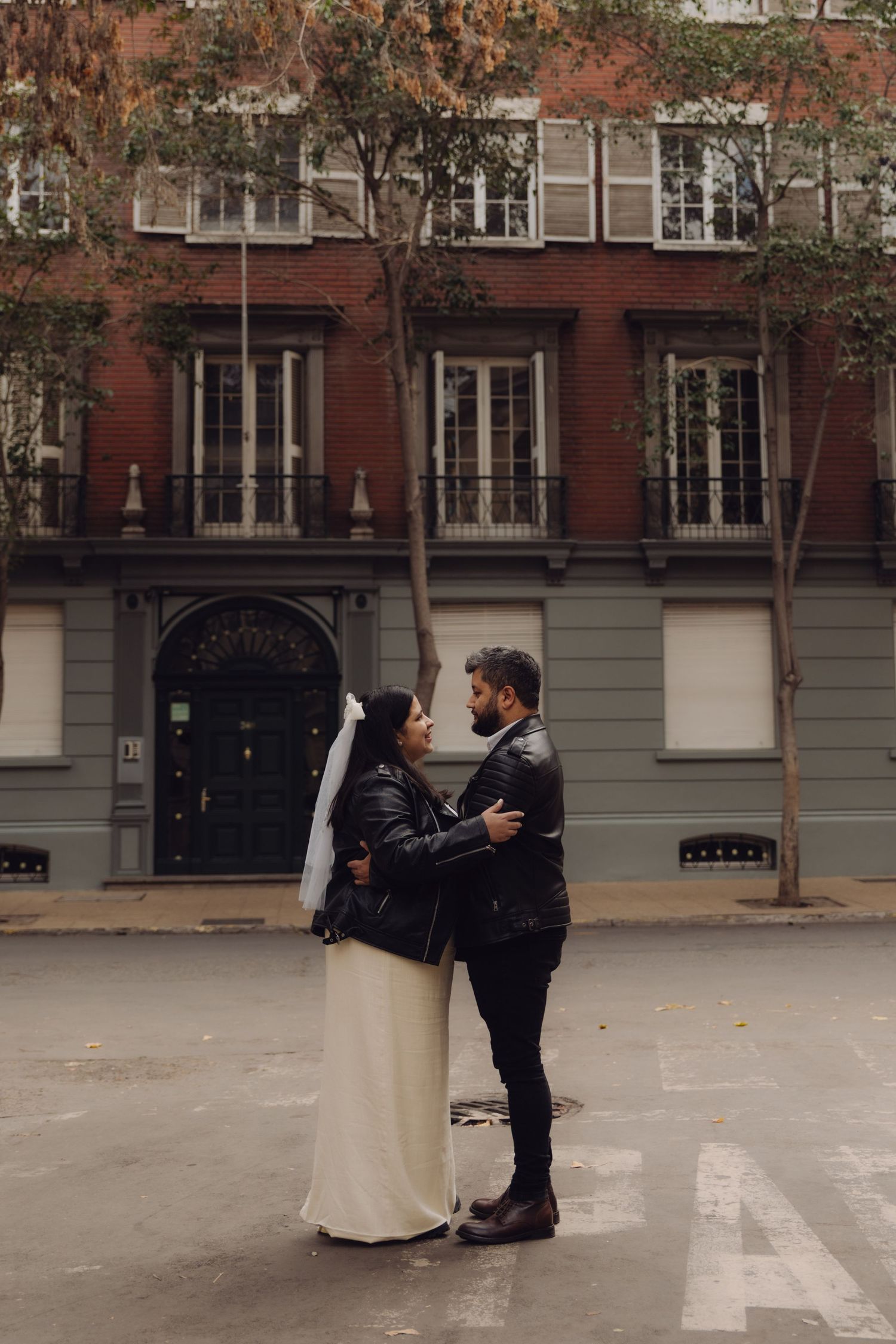 Bride and groom in leather jackets embrace in middle of wet city street with classic brick building behind them.