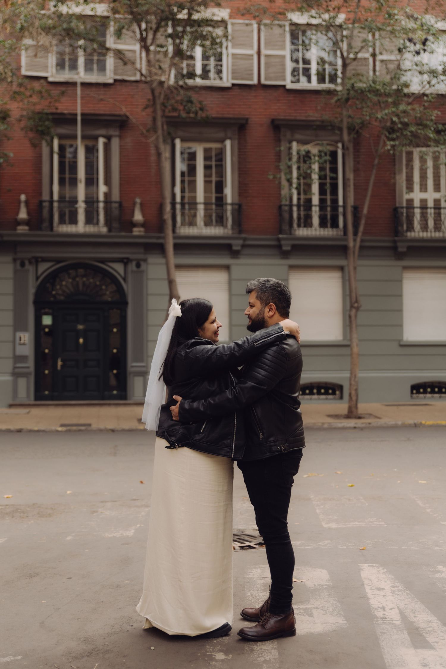 Bride in white dress with veil and groom in black leather jacket hold each other on urban street crosswalk.