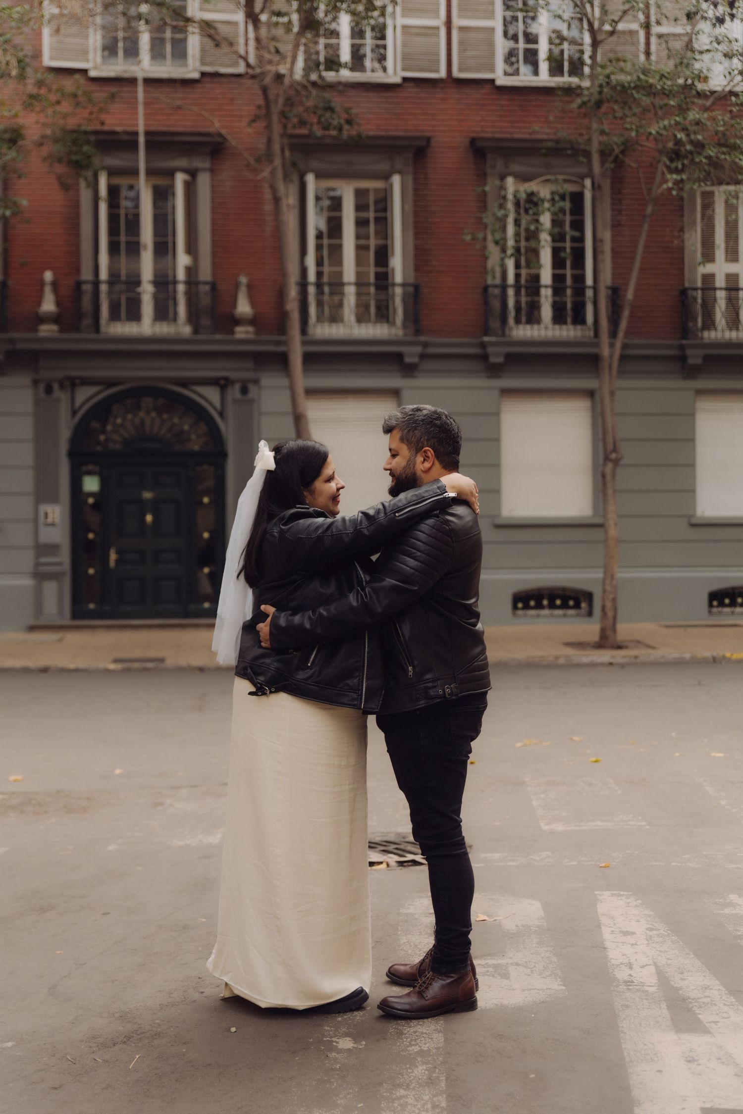 Bride in white dress and veil with groom in leather jacket embracing on urban street before red brick building.