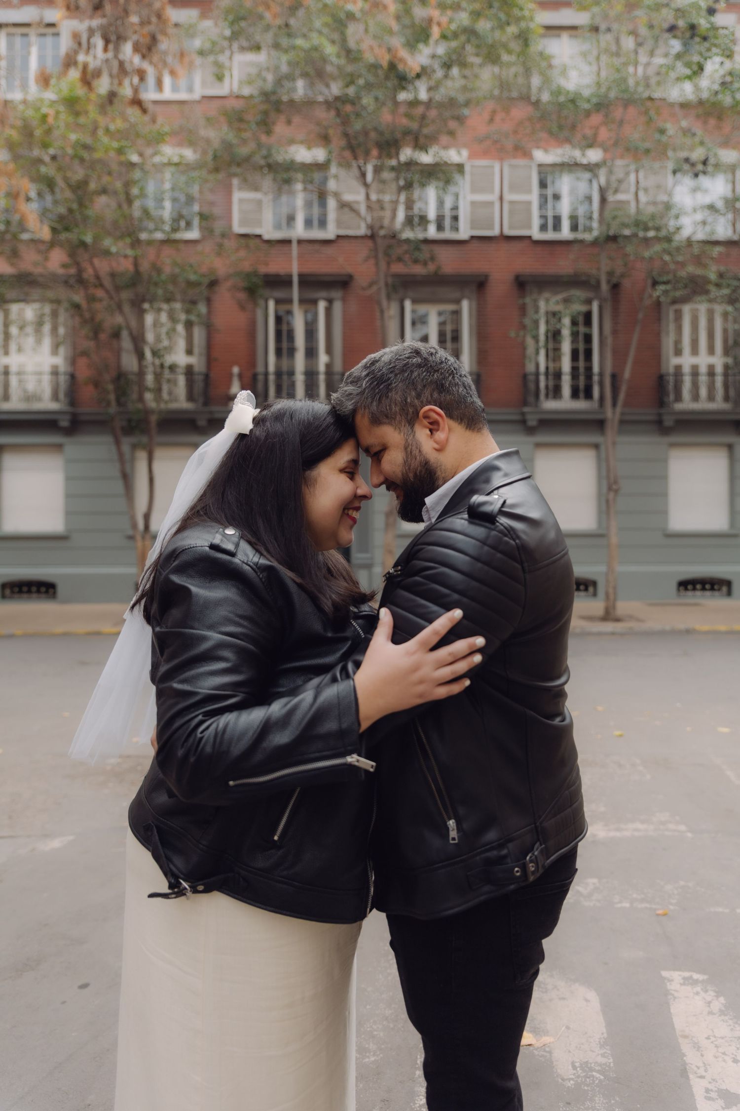 Newlyweds in matching black leather jackets touching foreheads romantically on city street with brick building backdrop.