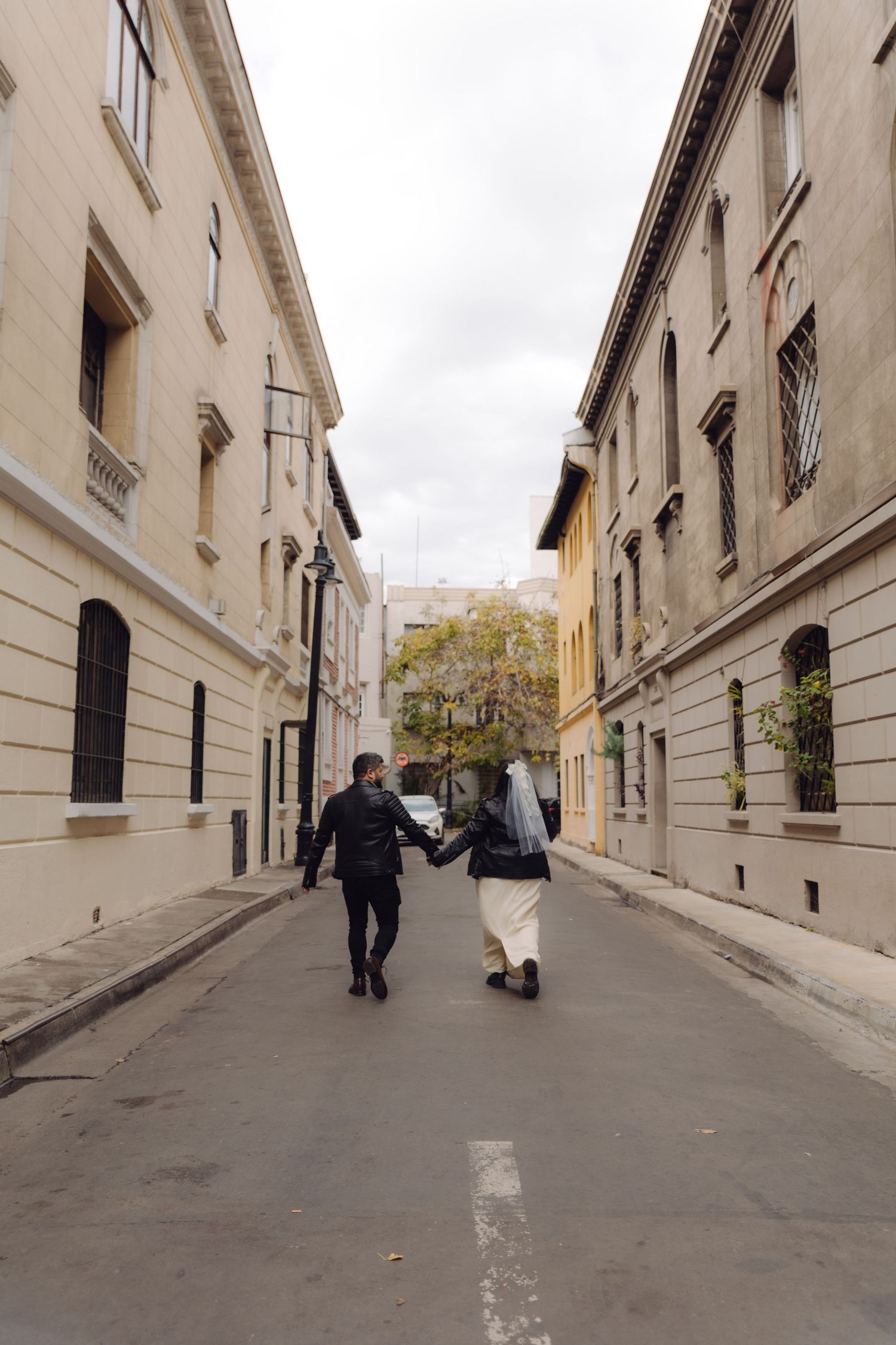 Bride and groom holding hands walking away down narrow European cobblestone street flanked by classical buildings.