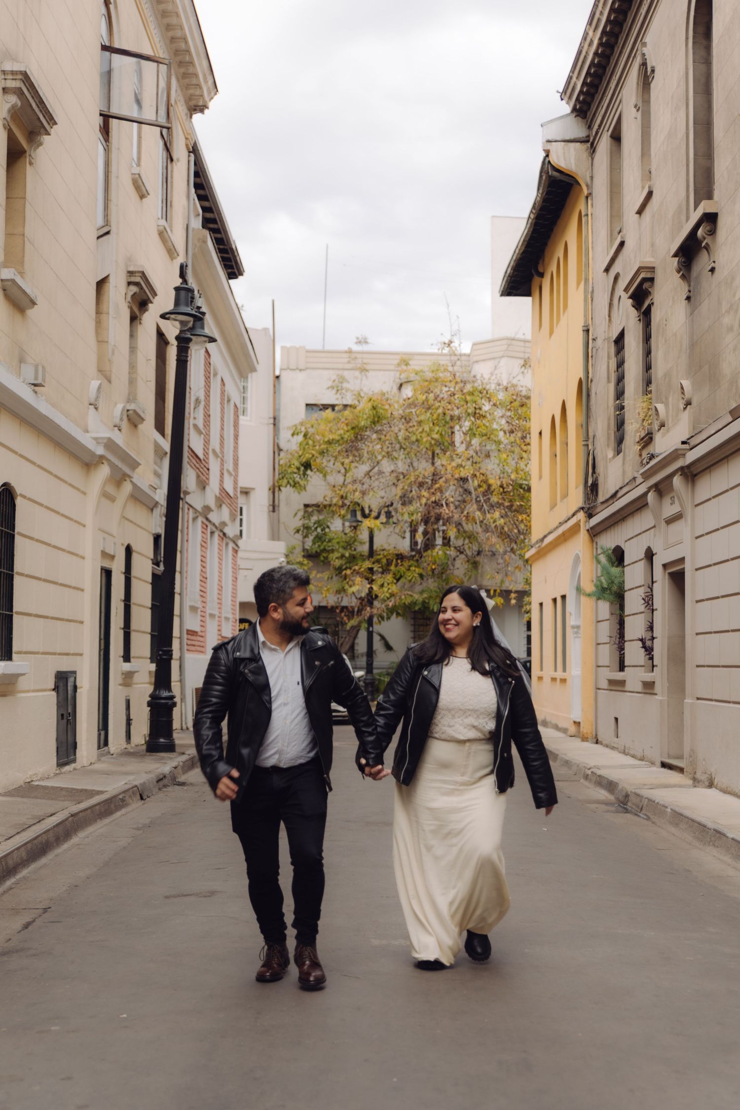 Happy couple in leather jackets holding hands walking through charming narrow European city street lined with buildings.