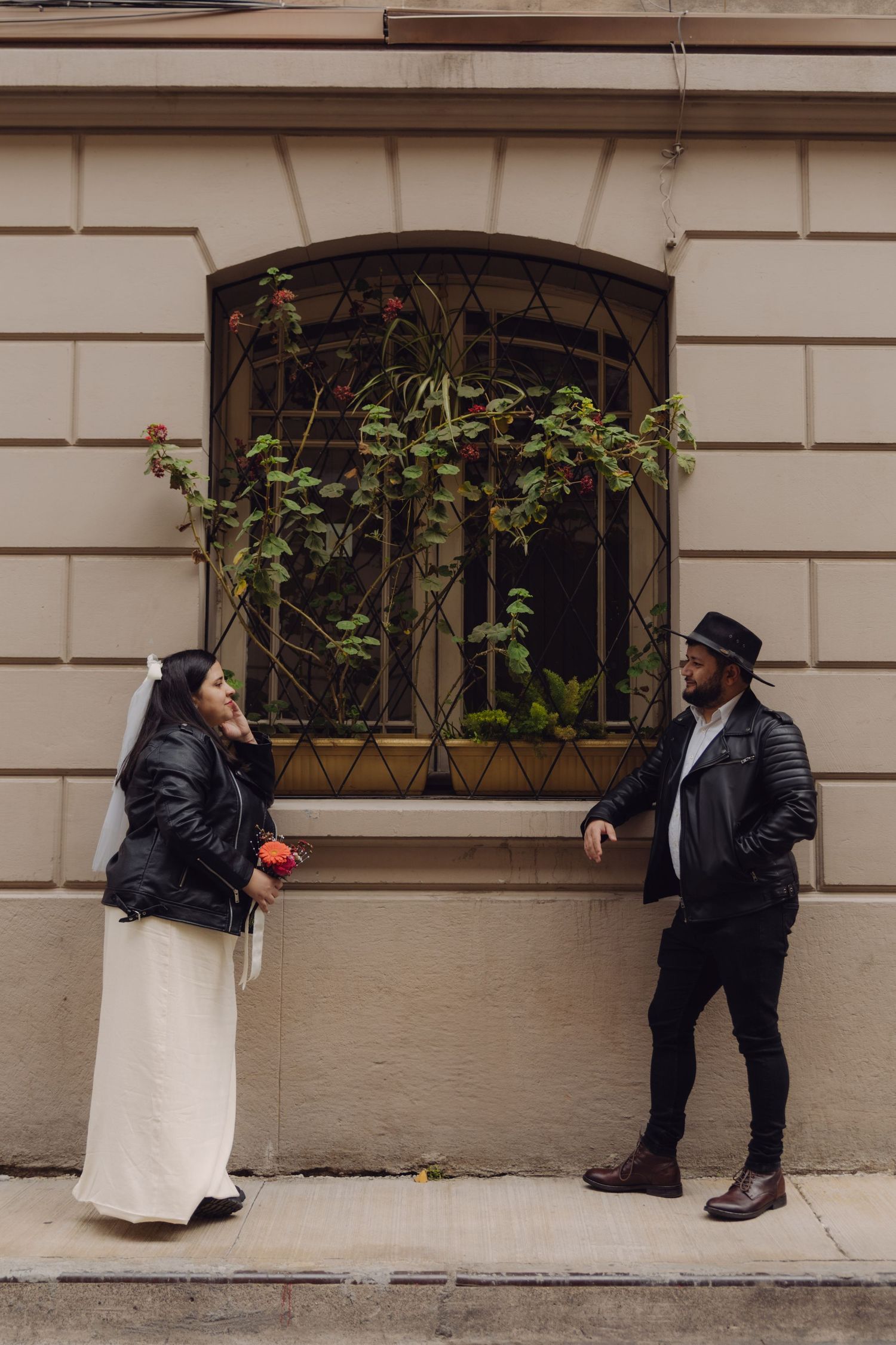 Bride with bouquet and groom in black hat standing apart flirtatiously by ornate barred window on city building.
