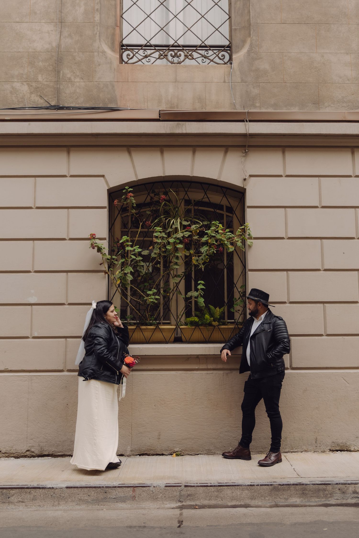 Wedding couple in leather jackets sharing playful moment beside arched barred window with climbing plants on building.