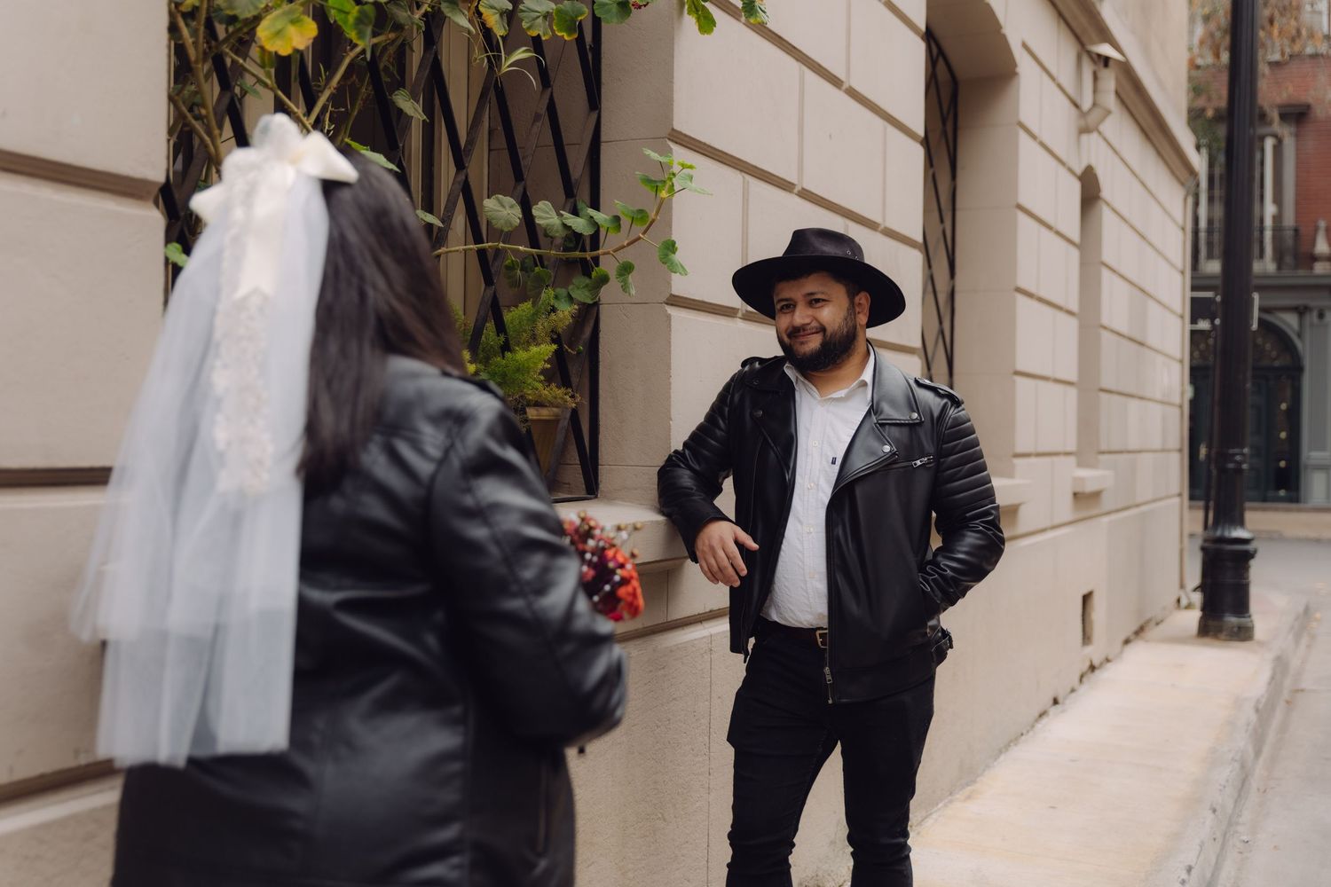 Groom in black hat and leather jacket smiling at veiled bride on charming European city sidewalk.