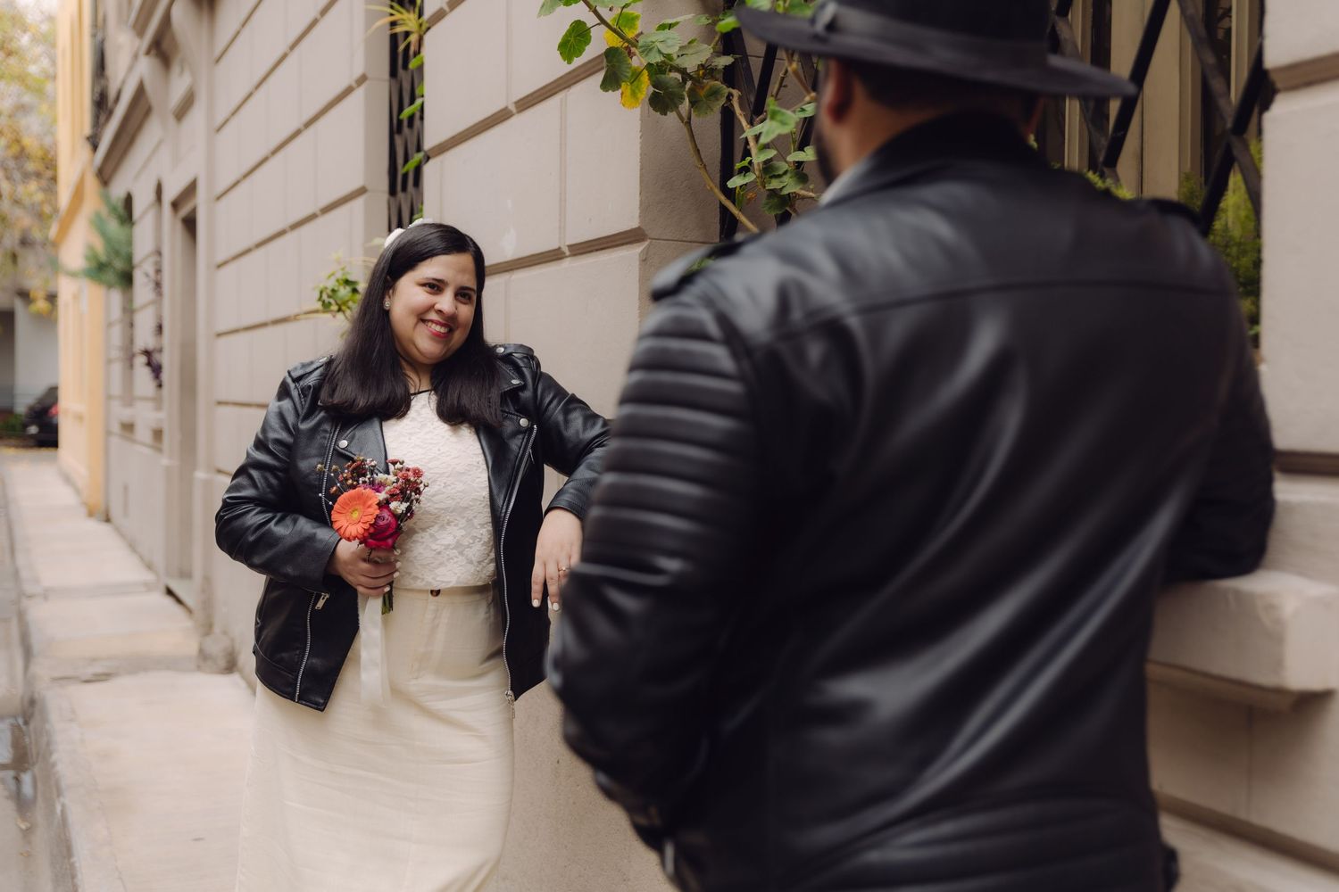 Smiling bride in leather jacket holding orange bouquet looking at groom on elegant urban sidewalk setting.