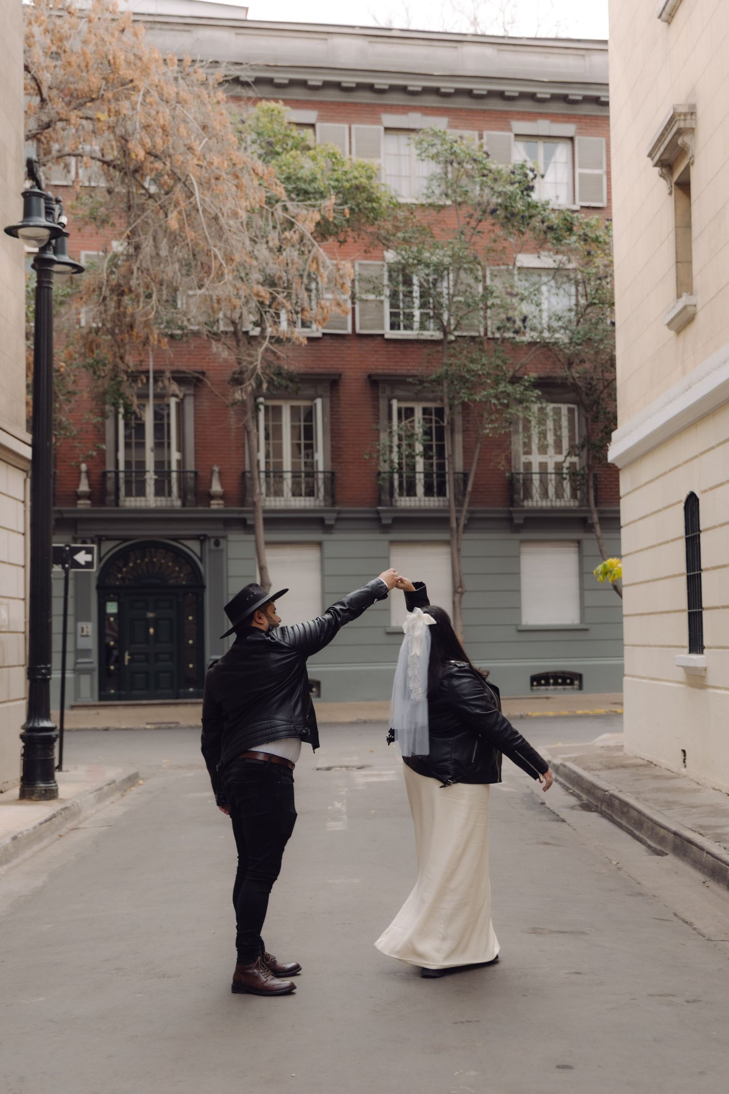 Groom in black hat spinning bride in white dress and leather jacket on charming European city street.