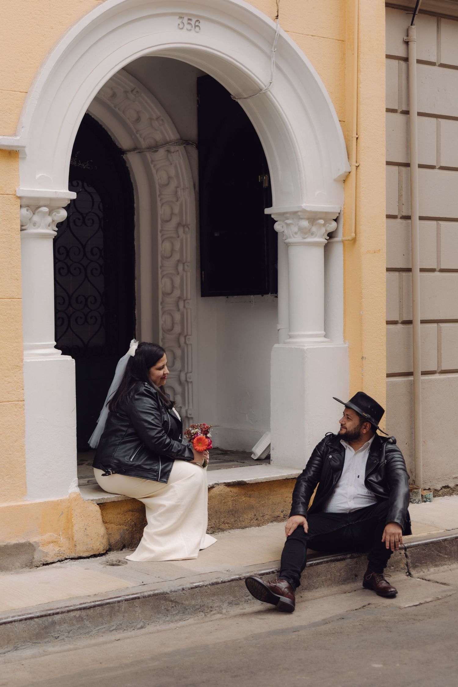 Bride and groom in leather jackets relaxing and chatting under ornate white arched doorway of yellow building.