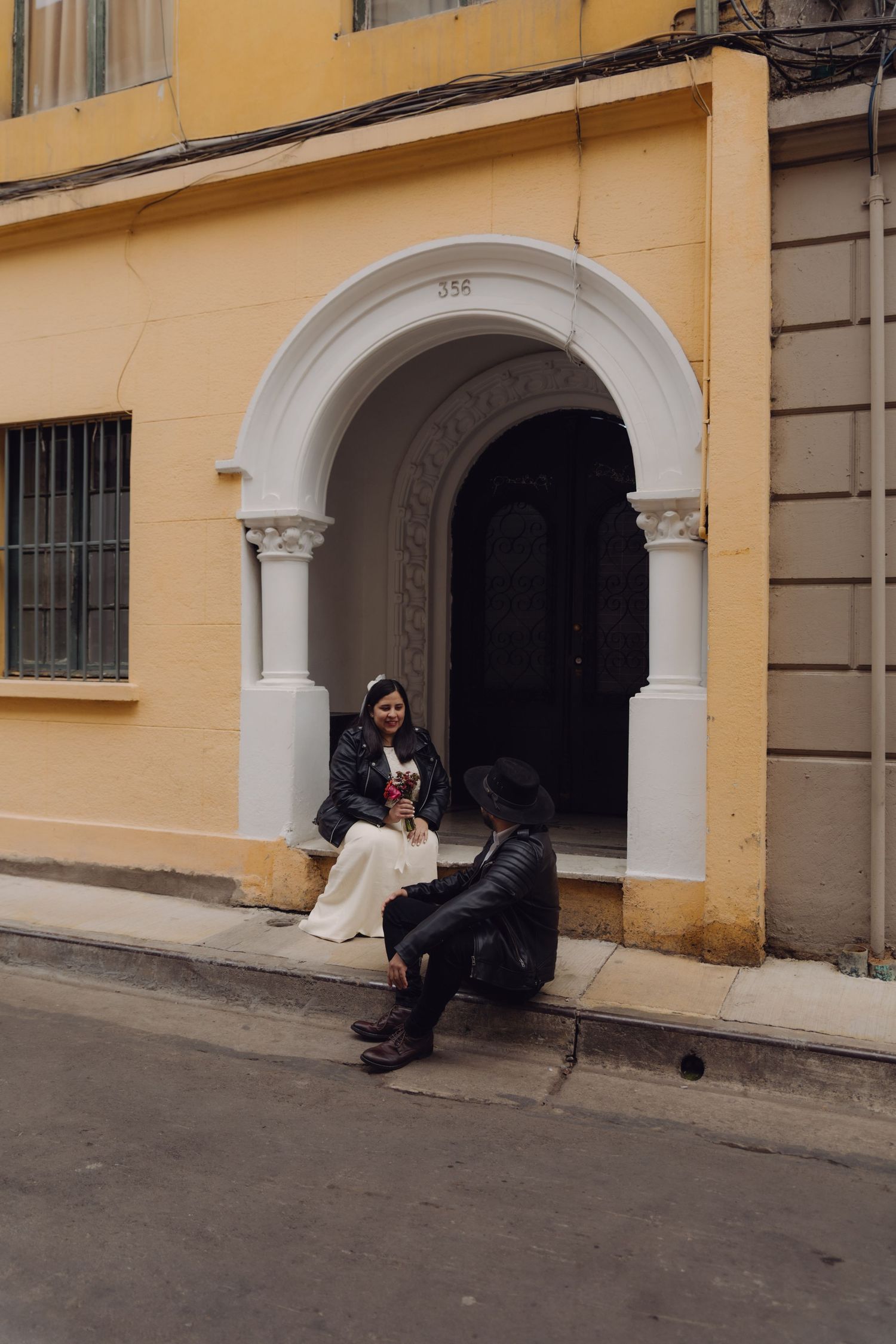 Couple sitting in a white arched doorway of a yellow colonial building on a city street.