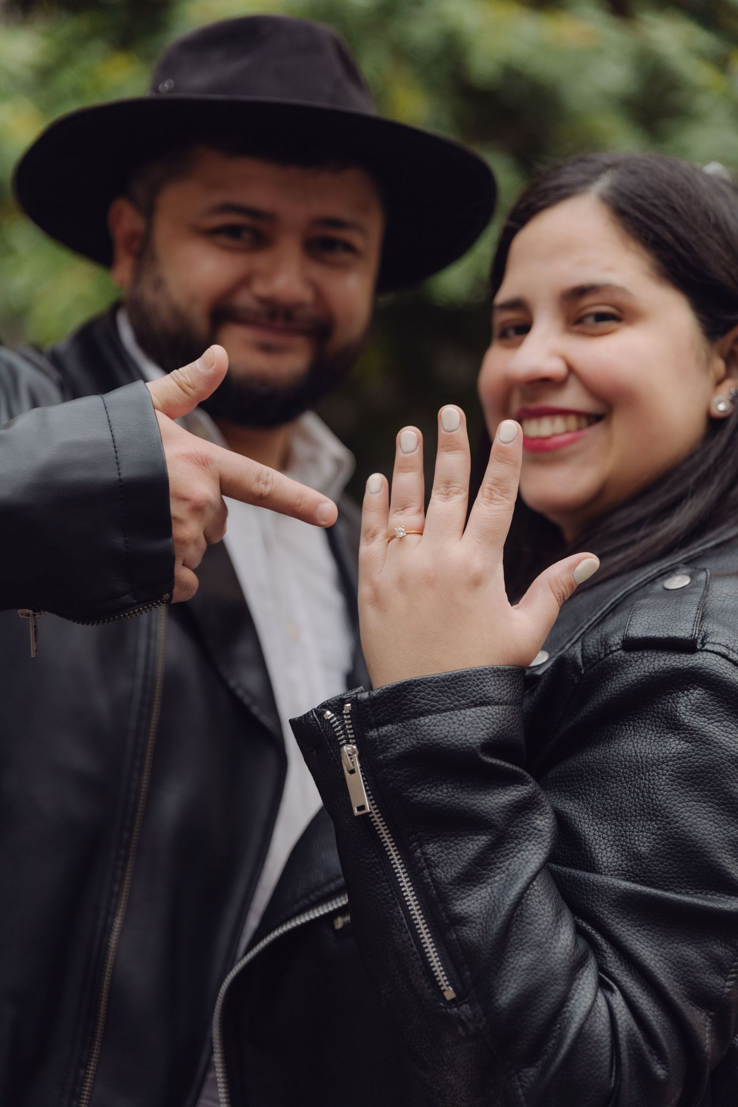 Engaged couple in leather jackets smiling and showing off engagement ring outdoors.
