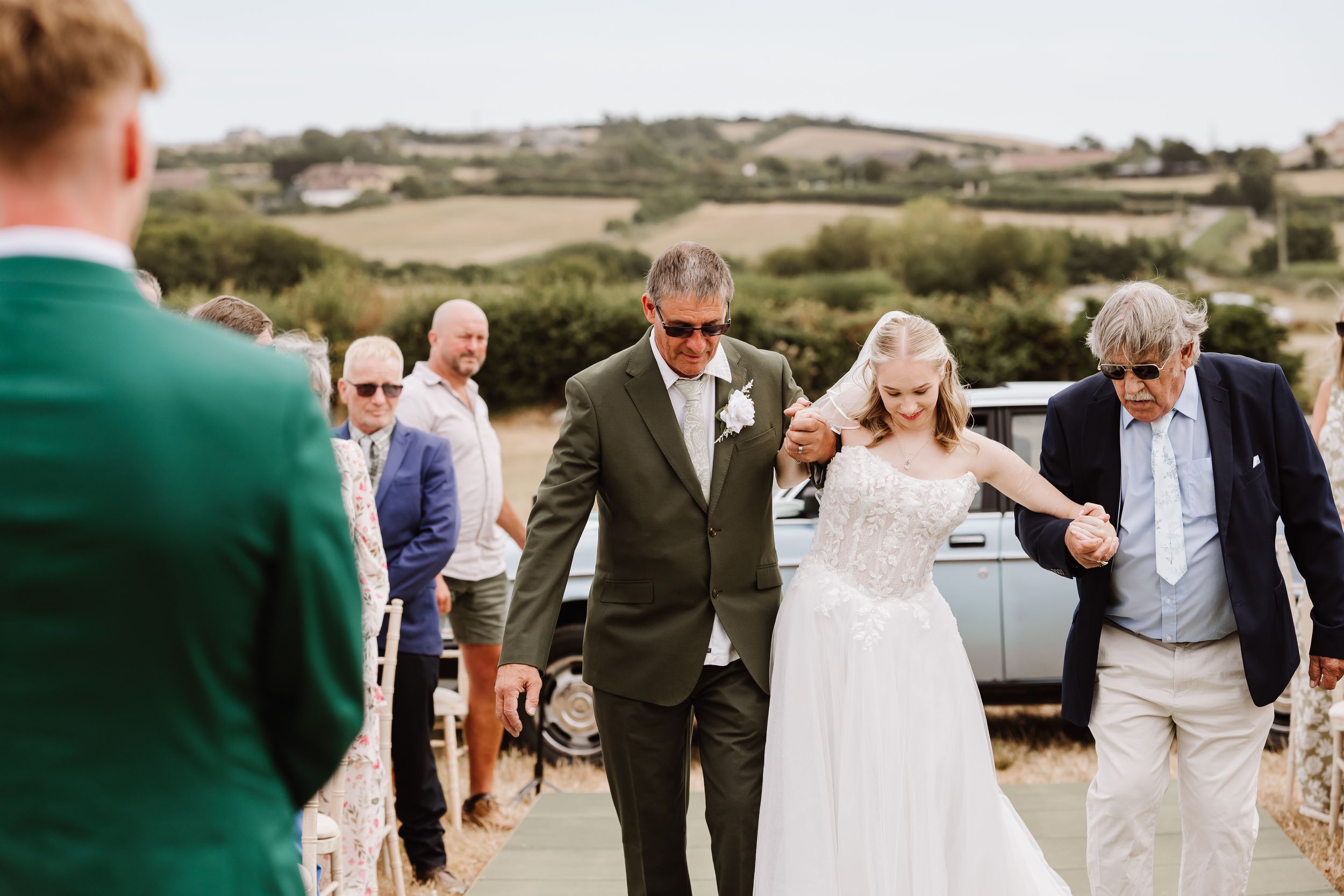 Bride in white gown escorted by two men in suits at outdoor wedding ceremony with guests in background at Moor Farm, Chickerell