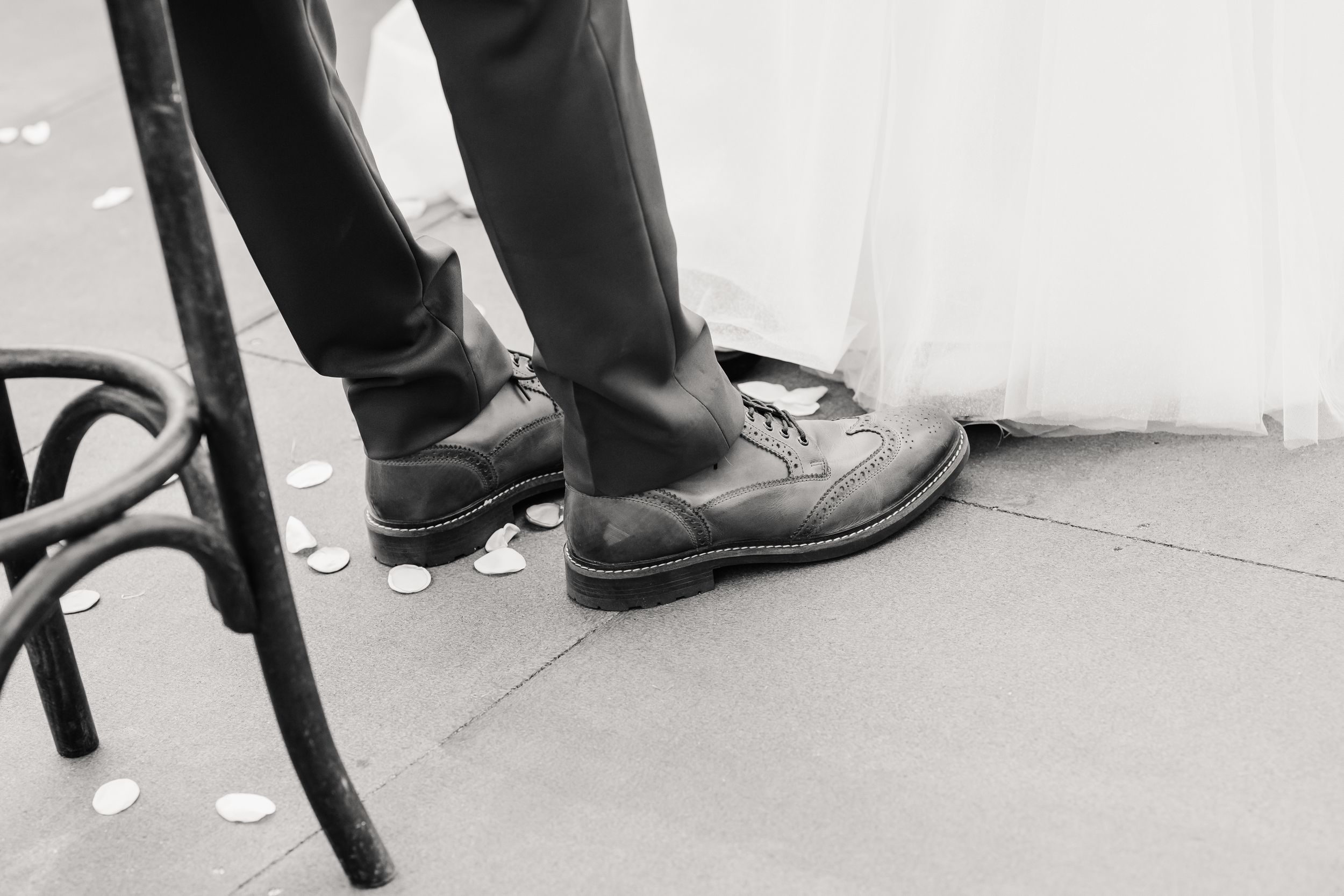 Black and white photo of groom's dress shoes and bride's wedding gown hem on stone floor with scattered petals.