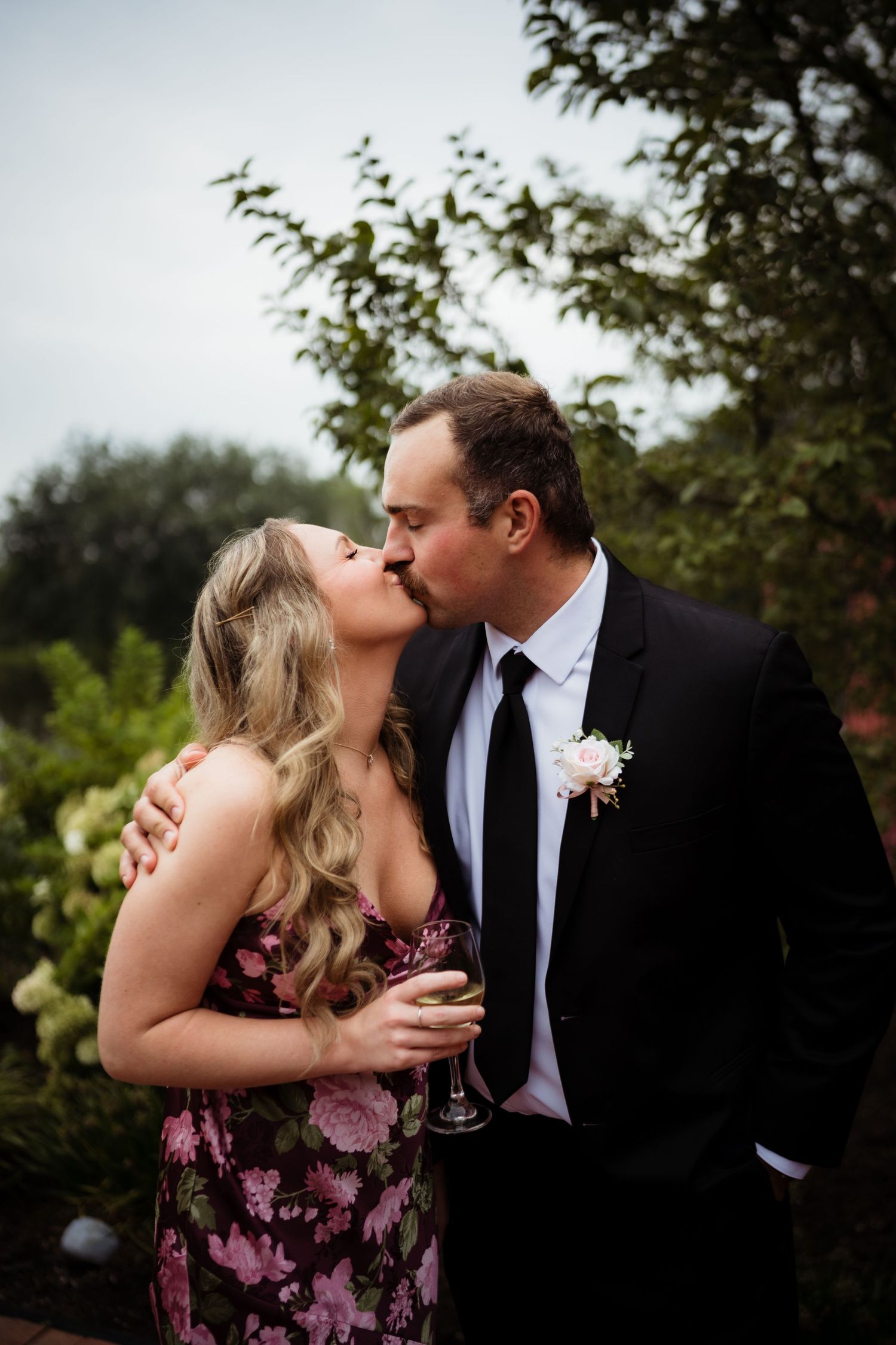 A couple shares a romantic kiss outdoors, man in black suit with boutonniere, woman in floral dress holding champagne glass.