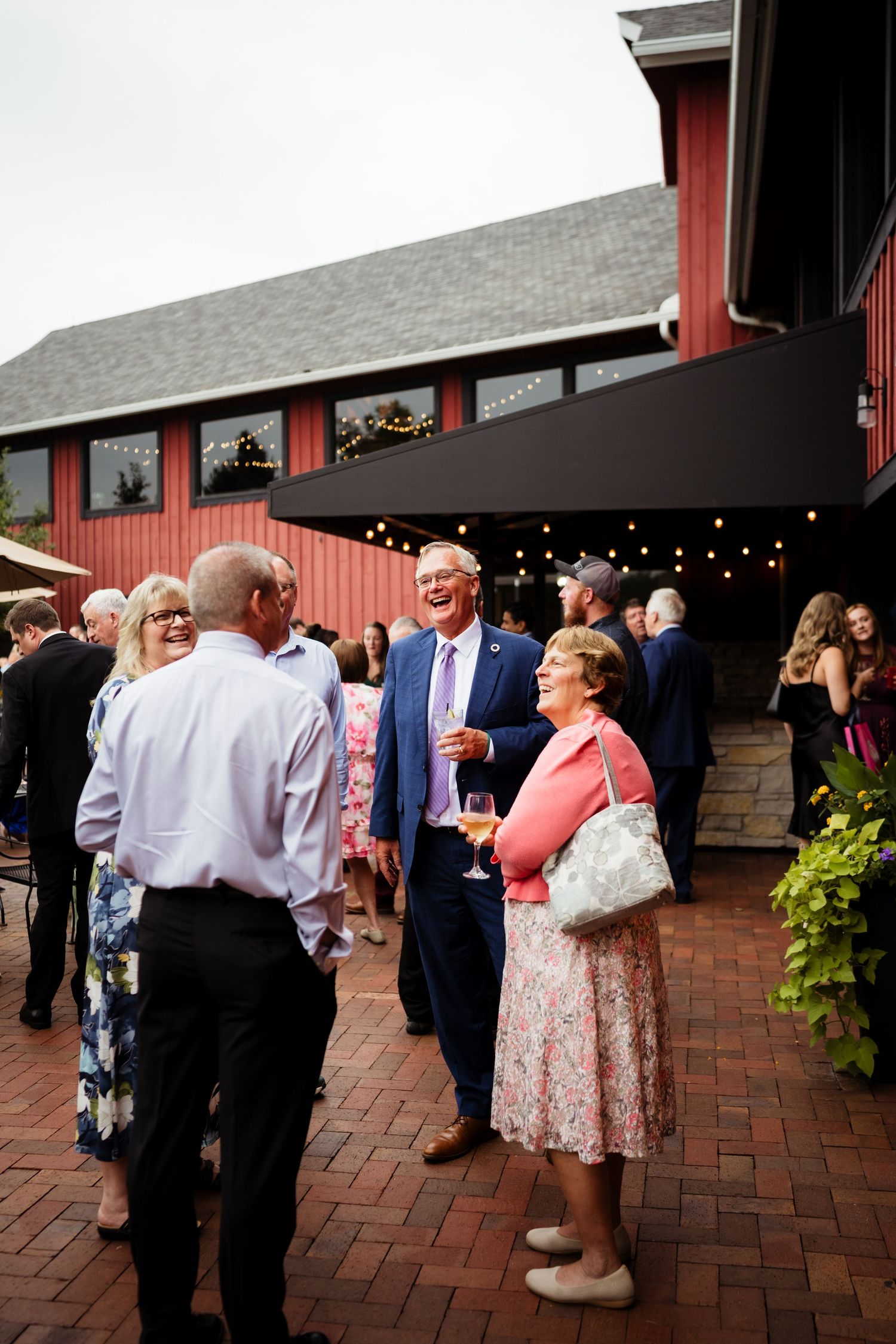 Wedding guests socializing outdoors on brick patio near red barn venue with string lights.