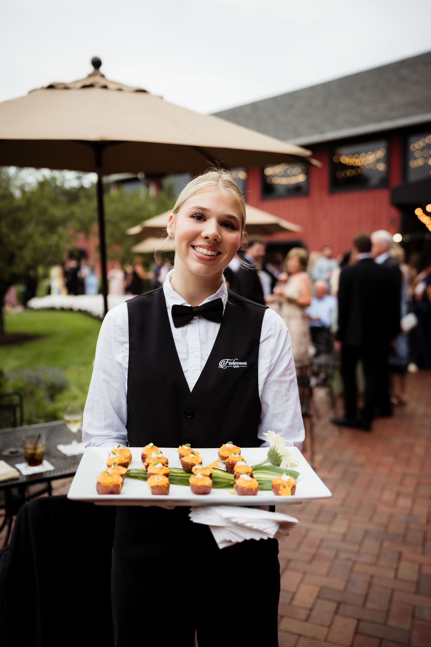 Smiling waitress in bow tie holds tray of appetizers at outdoor catered event near red barn venue.