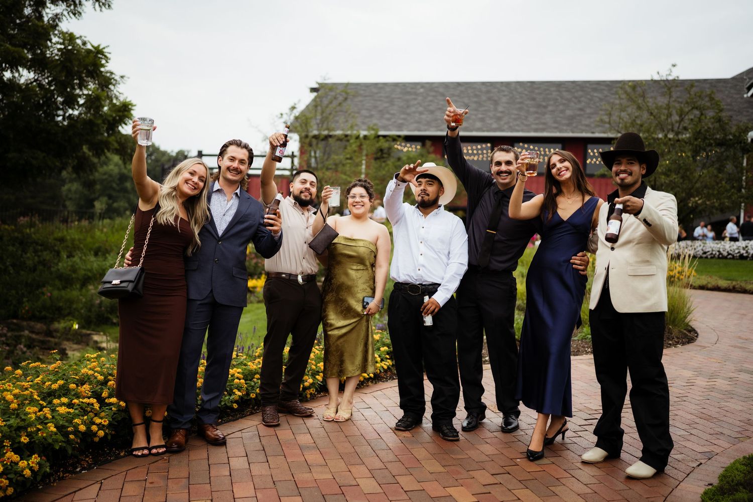 Group of elegantly dressed wedding guests celebrating outdoors on a brick pathway surrounded by lush garden greenery.