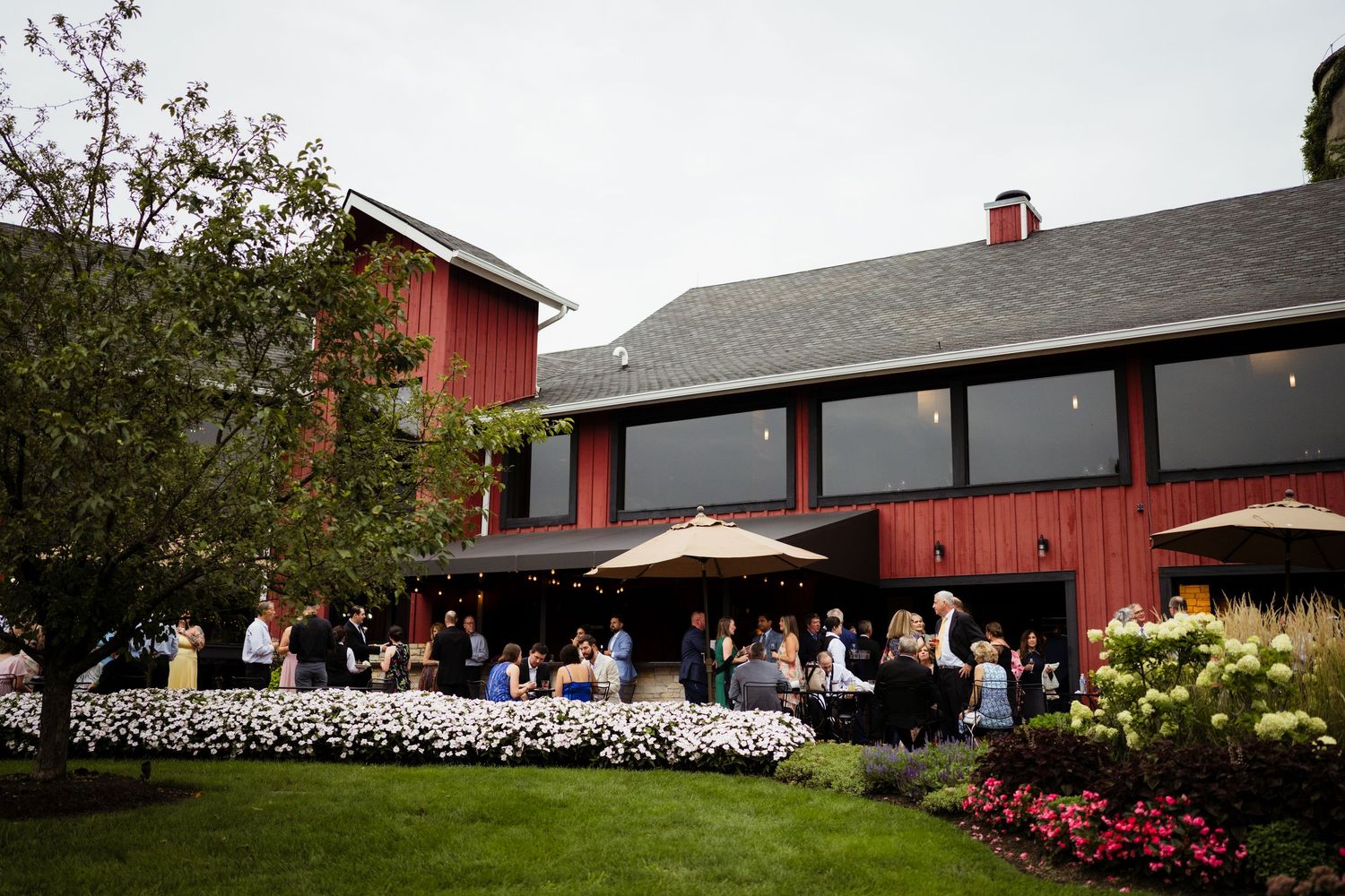 Red barn wedding venue exterior with guests mingling on manicured lawn surrounded by lush gardens.