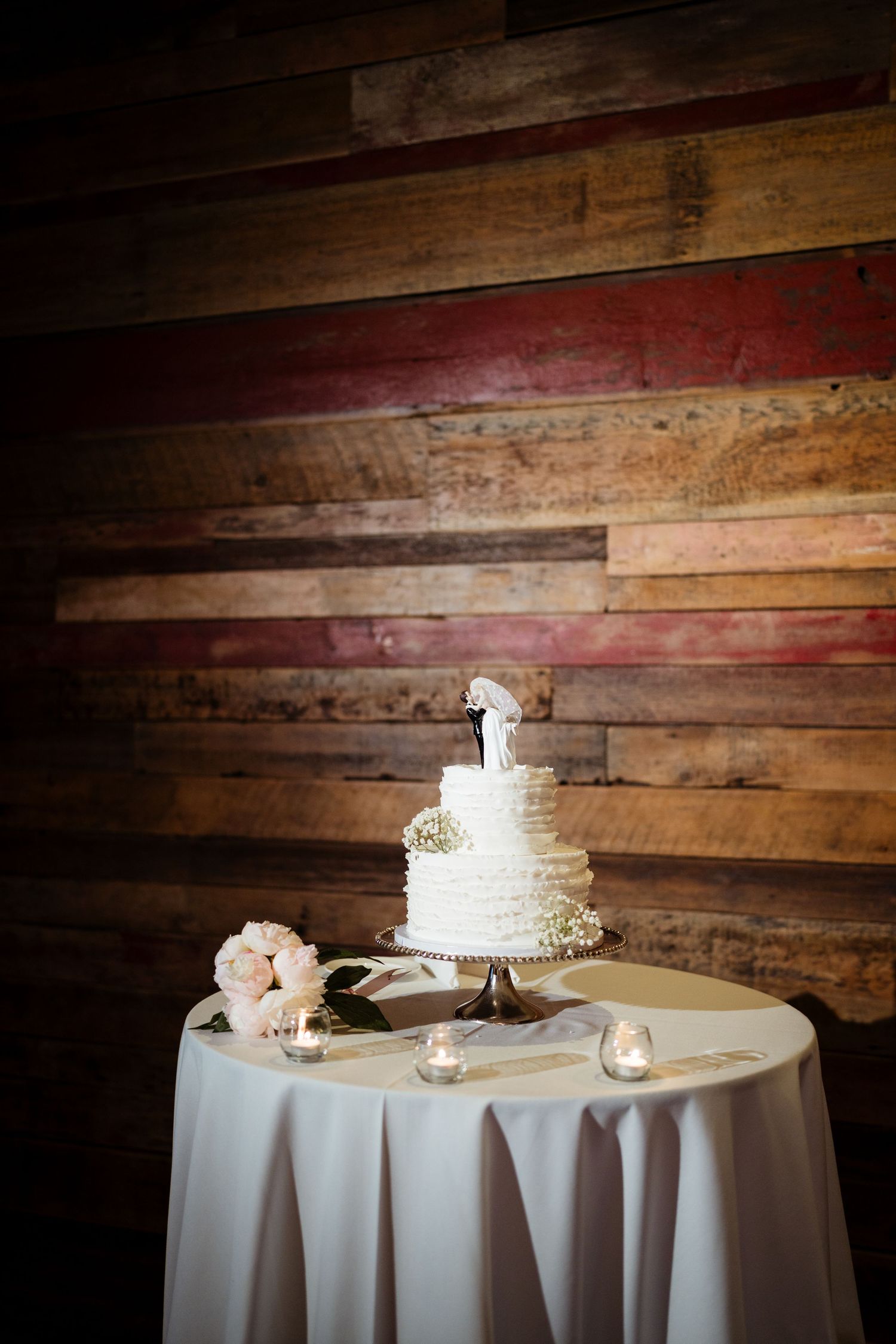 Three-tier white wedding cake with bride and groom topper on draped table against rustic reclaimed wood wall.