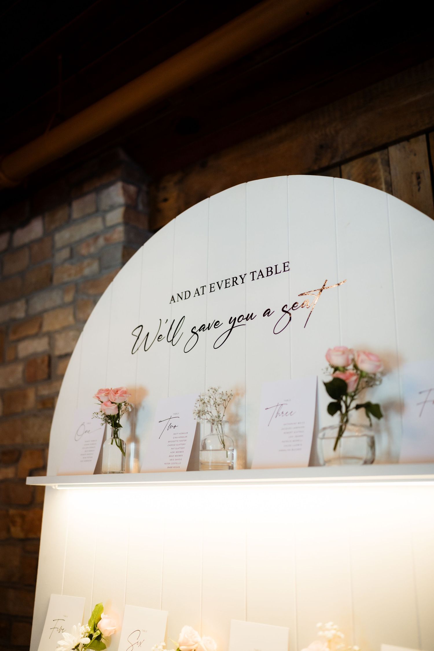 White arch wedding seating chart display with script text and pink rose floral accents on brick wall.