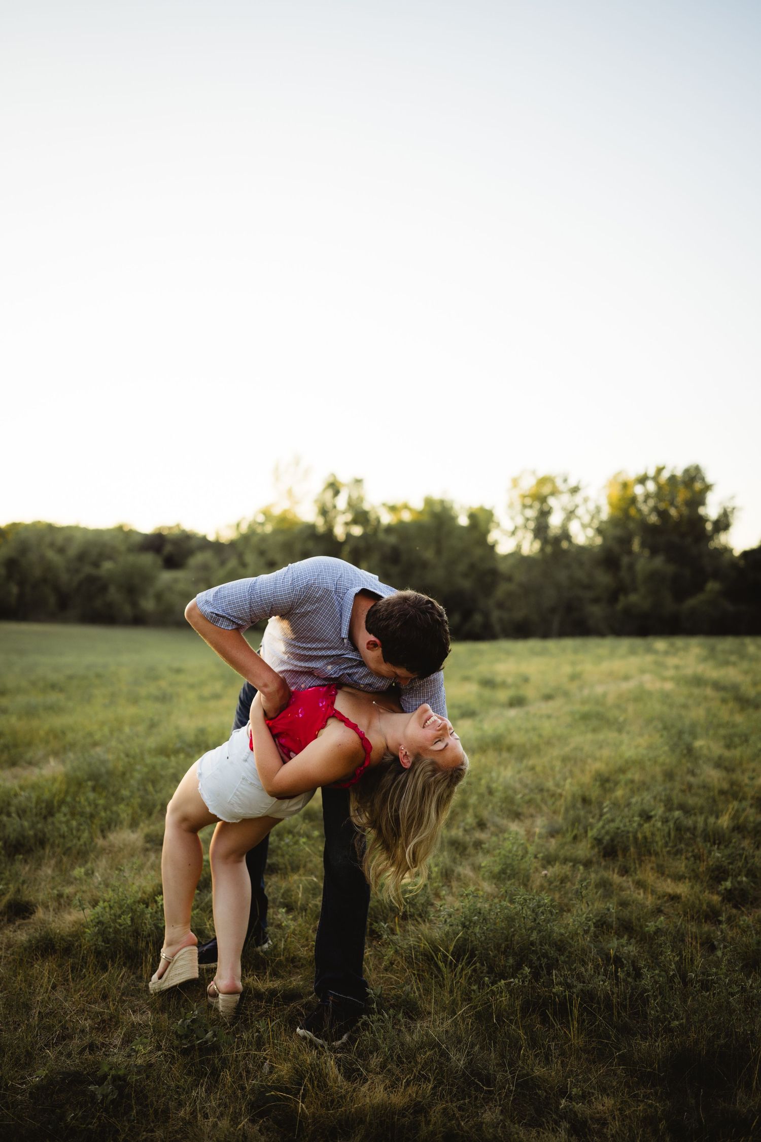 Couple sharing a romantic dip kiss in a lush green field at sunset, man in gray shirt, woman in red top.