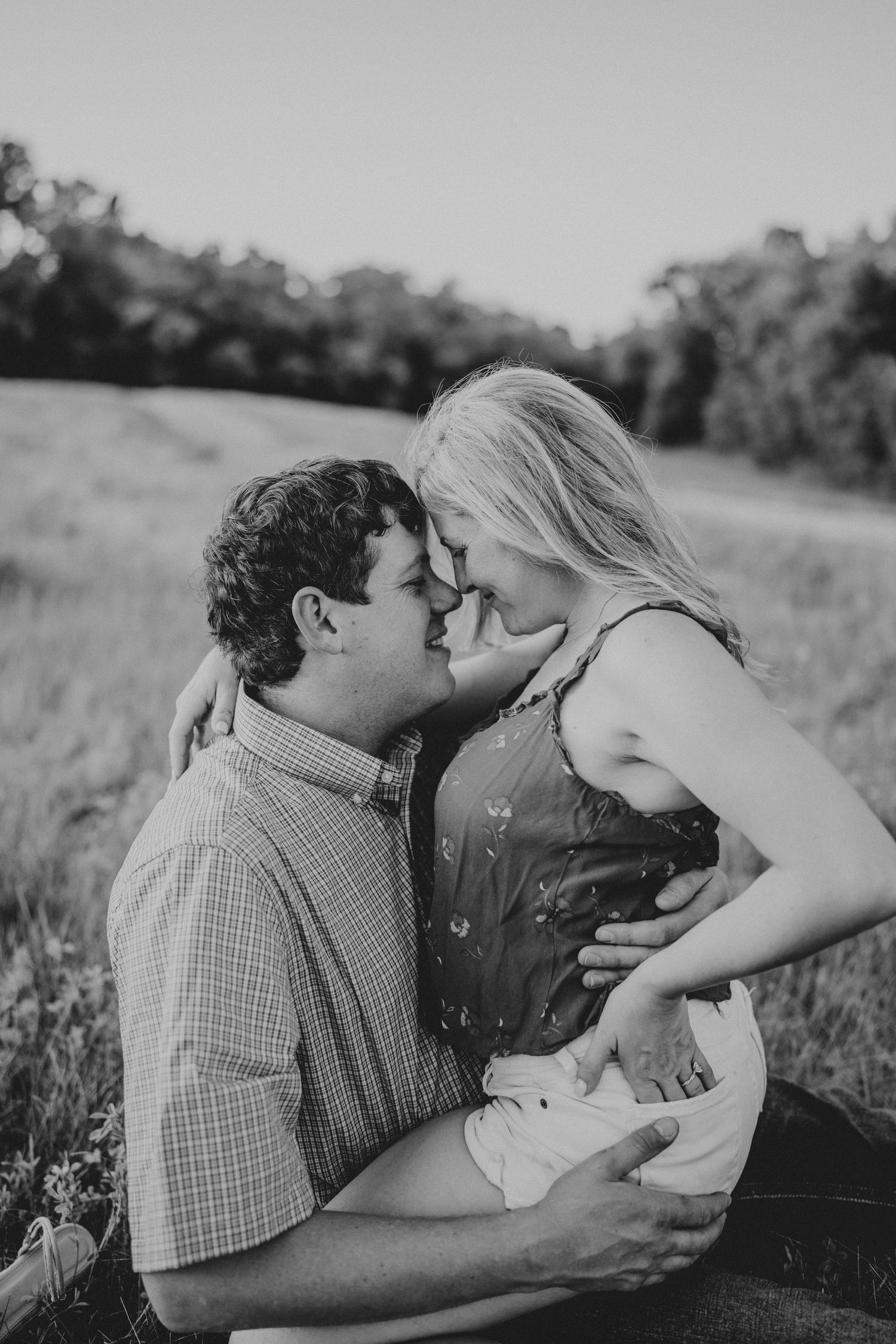 Black and white photo of a couple sharing a tender moment, almost kissing in a sunlit grassy field.