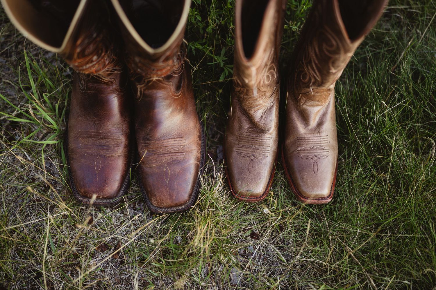 Two pairs of leather cowboy boots standing on green grass, one dark brown and one tan with decorative stitching.