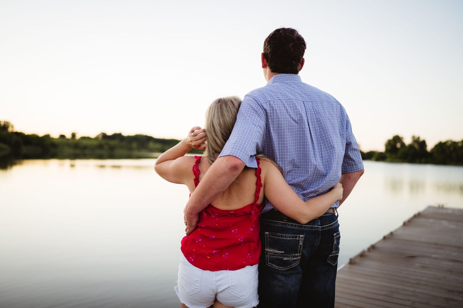 Couple embracing on a wooden dock overlooking a calm lake at sunset, woman in red top and white shorts.