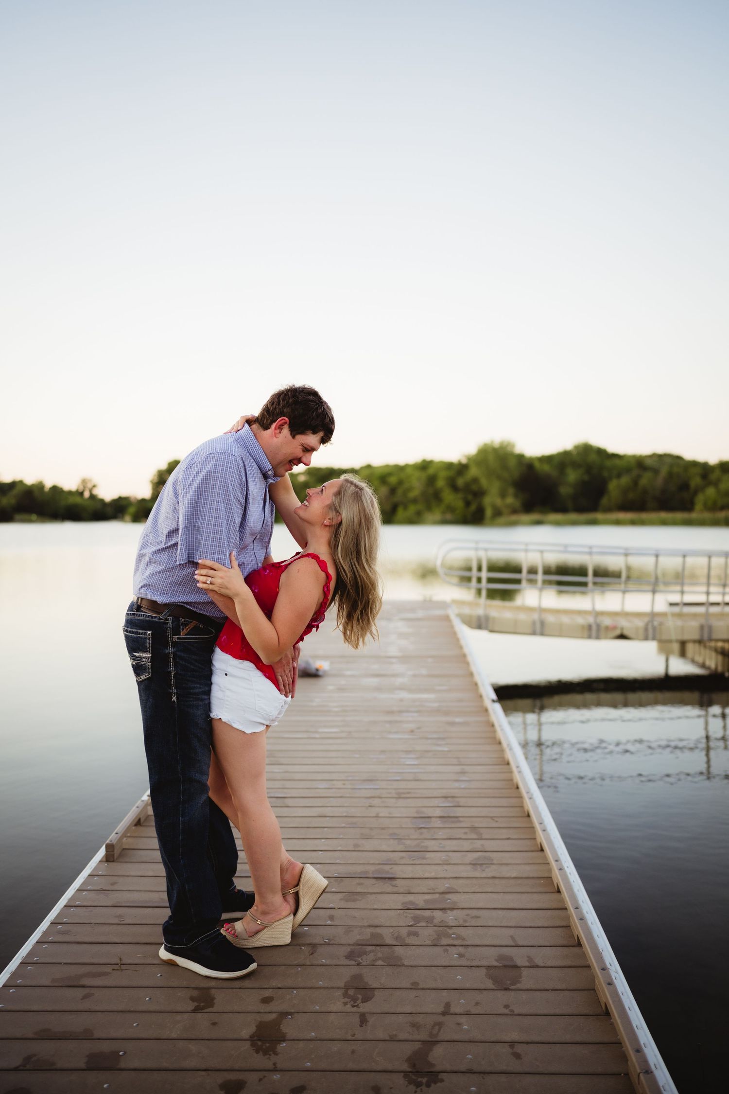 Couple sharing a romantic kiss on a wooden dock by a calm lake at sunset, woman in red top and white shorts.