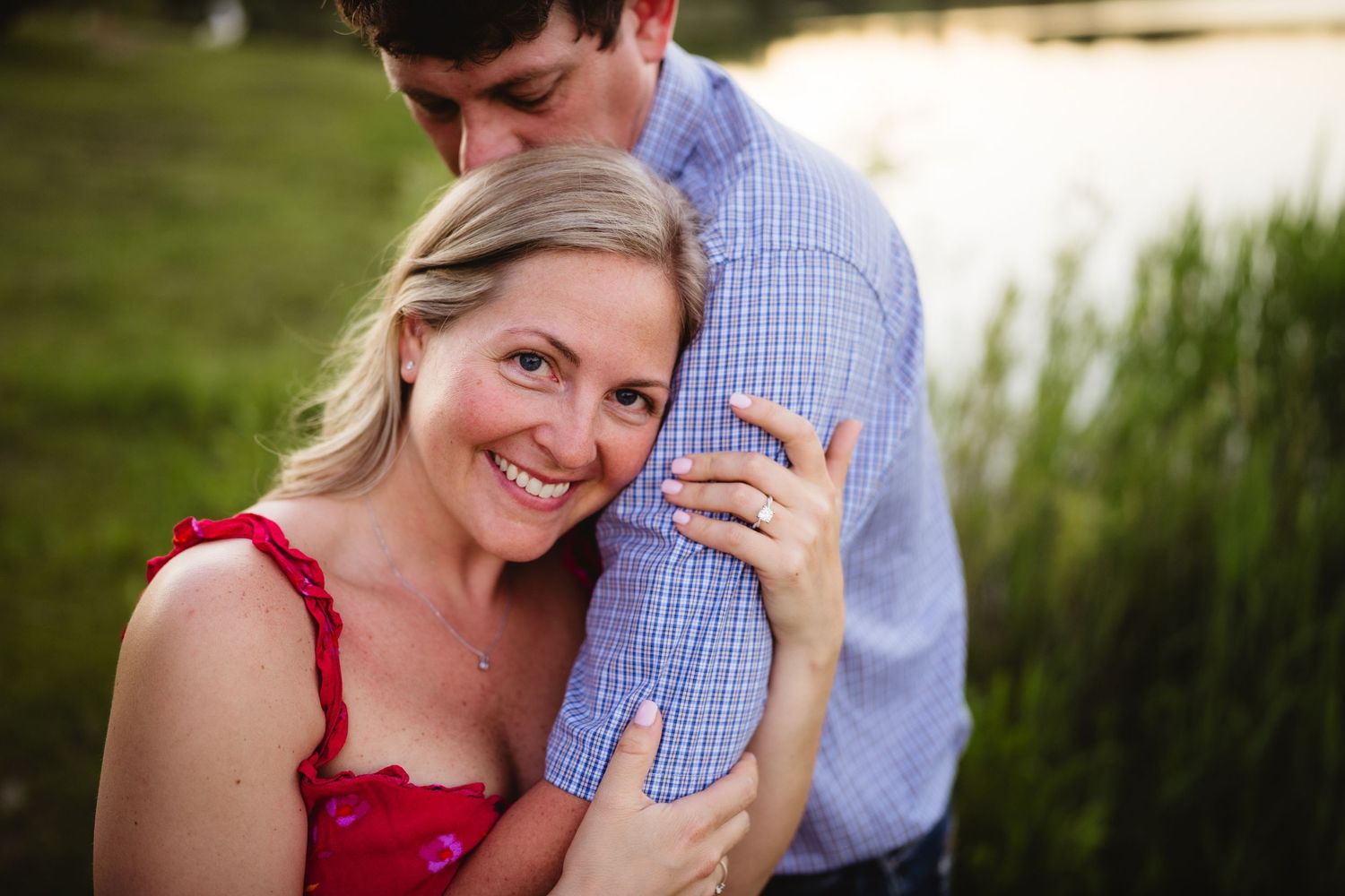 A smiling blonde woman in a red dress hugs a man in a blue shirt near a pond at sunset.