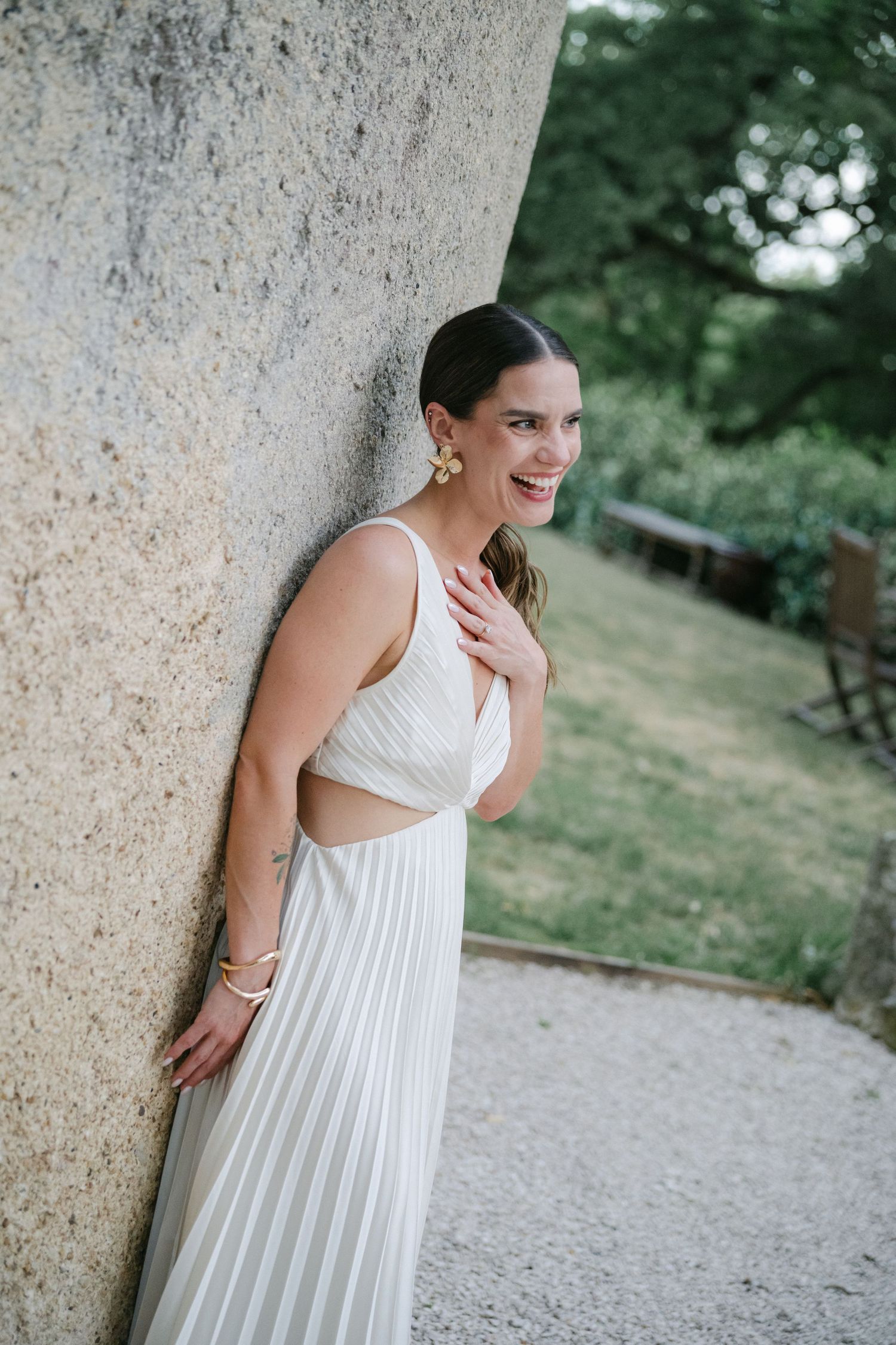 A laughing bride and groom in wedding attire share a joyful moment on a vine-covered balcony beside a rustic terracotta urn.