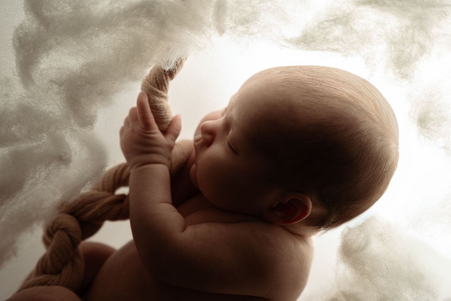 Newborn baby curled in fetal position with umbilical cord, sleeping peacefully against soft cloudy background.
