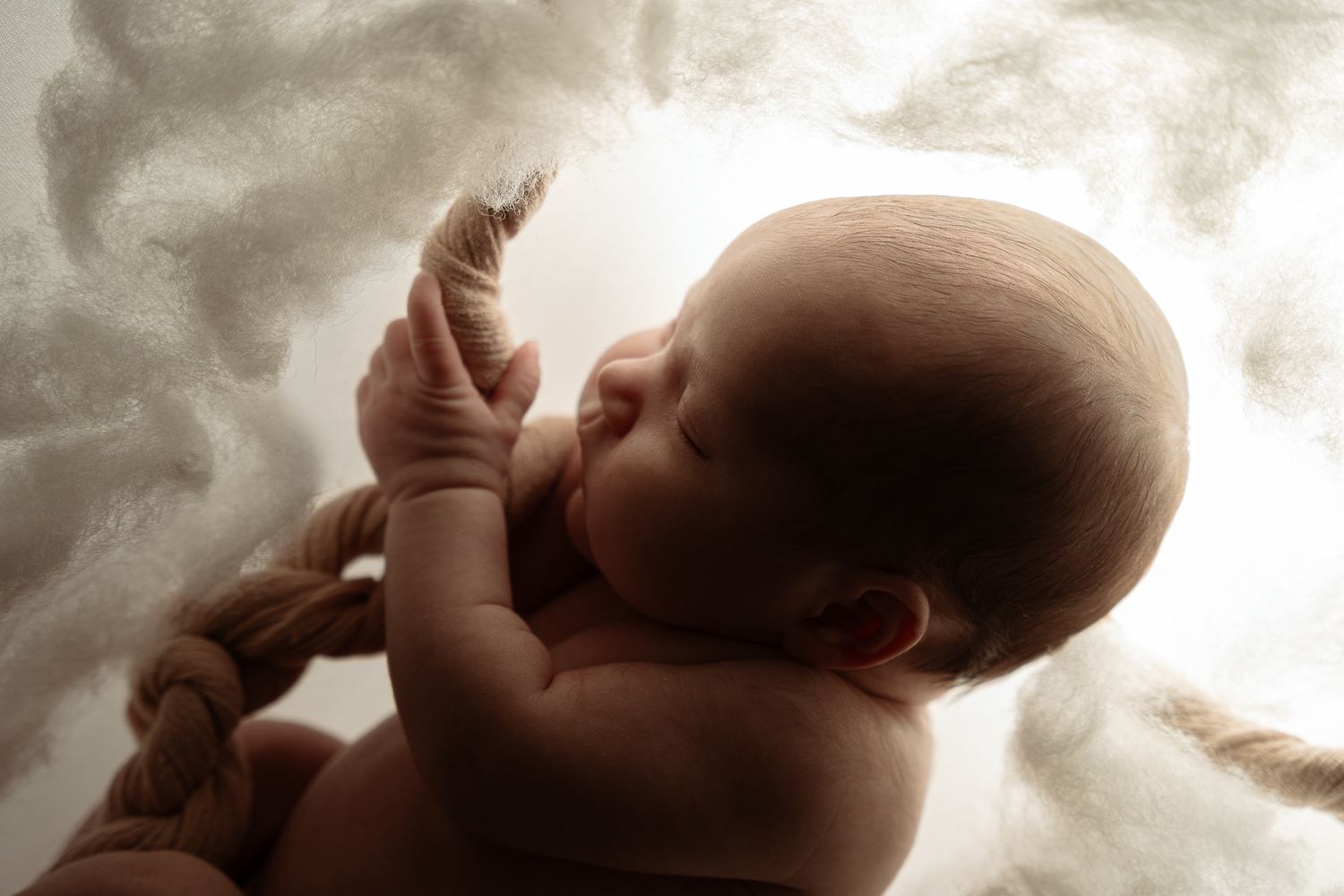 Newborn baby curled in fetal position with umbilical cord, sleeping peacefully against soft cloudy background.