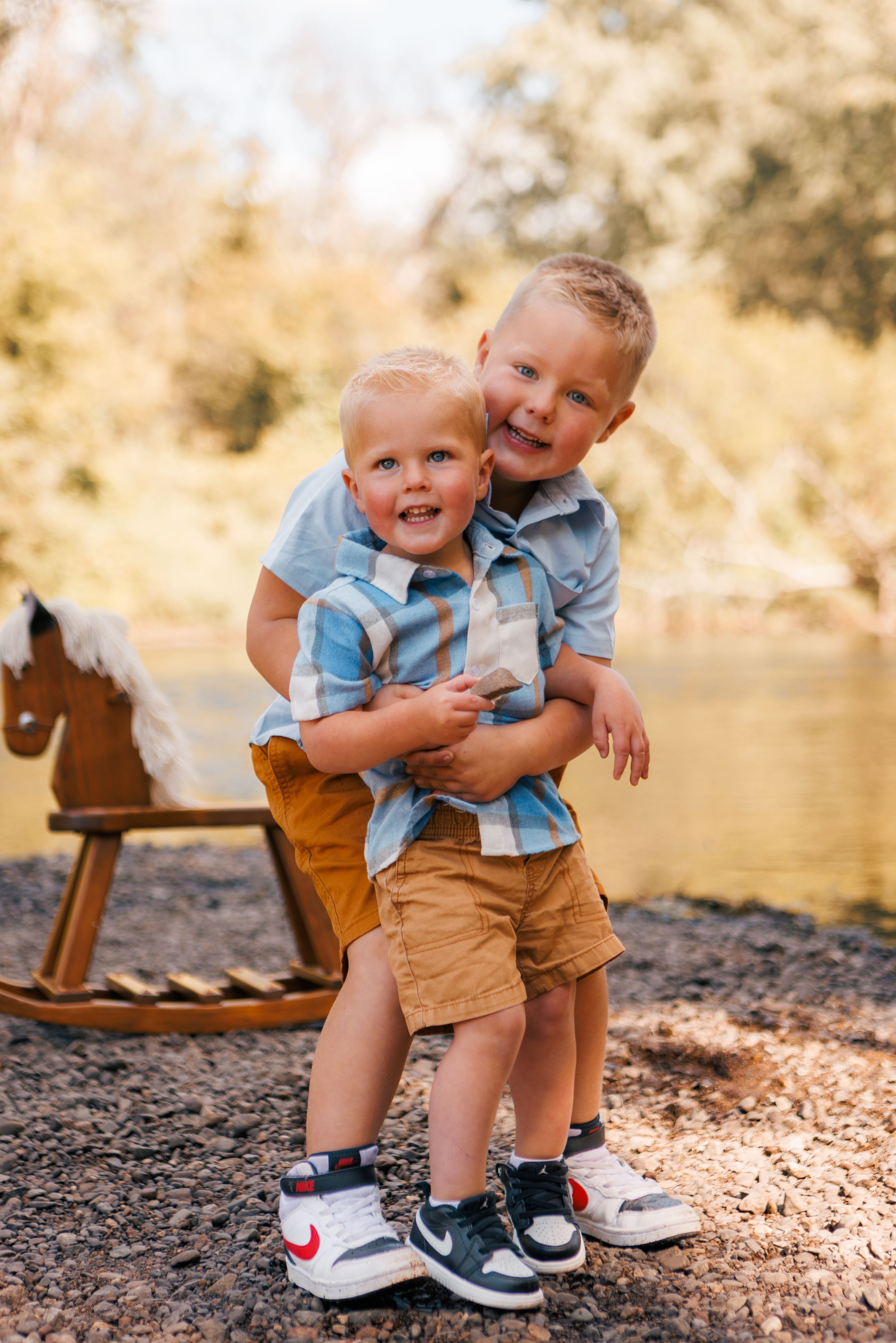 Two young brothers hugging and smiling outdoors near a wooden rocking horse toy with trees in background.