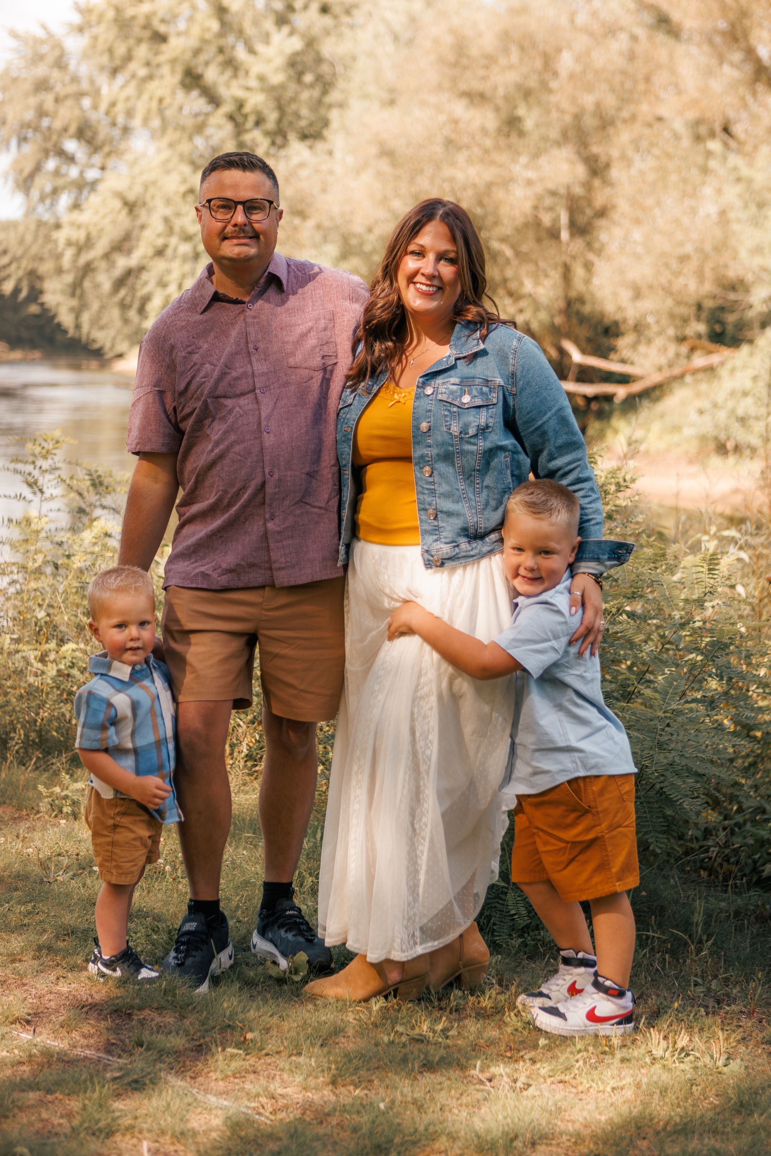 Family of four posing outdoors near trees, parents with two young boys in coordinated casual outfits, smiling for a portrait.